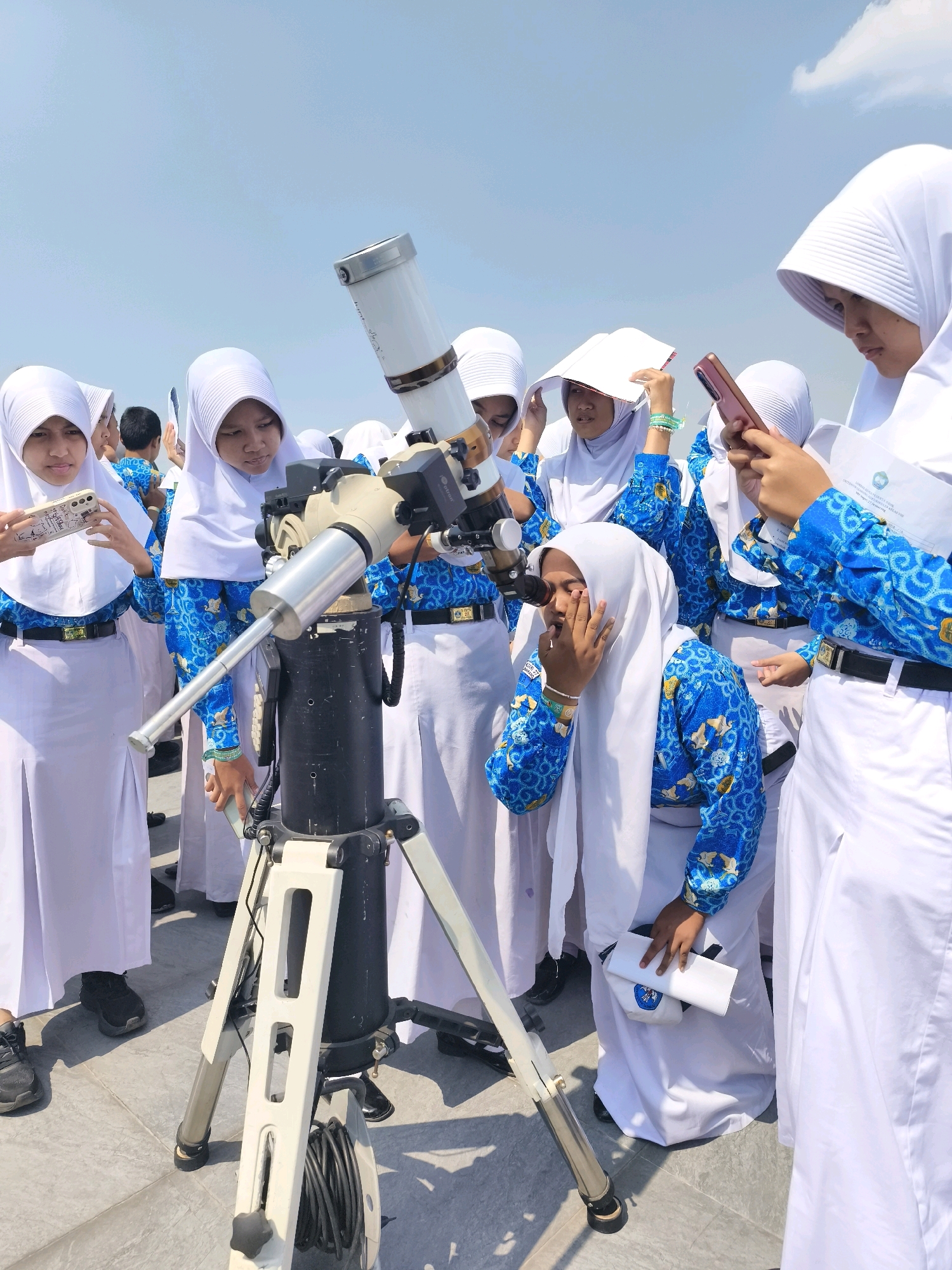 The image shows a group of young women, likely students, dressed in white hijabs and blue and white patterned school uniforms, gathered around a telescope. The scene appears to be outdoors, with a clear blue sky in the background, suggesting a bright, sunny day. One woman is looking through the telescope, while others are holding cell phones, a book, and a document. The telescope is mounted on a tripod, with cables coiled at its base. The ground appears to be paved with tiles. The women's attire and the presence of a school document suggest an educational activity. The overall impression is one of curiosity and learning, with the students engaged in observing something through the telescope.
The question asks for the location in the city of Semarang, Indonesia. Based on the visual information alone, it is not possible to pinpoint the exact location in Semarang. However, the presence of a school group and an outdoor observatory setup might suggest a location such as a school campus with an observatory, a science center, or a public park equipped for astronomical observation. Without further context or identifying landmarks, it's impossible to be more specific about the location in Semarang.