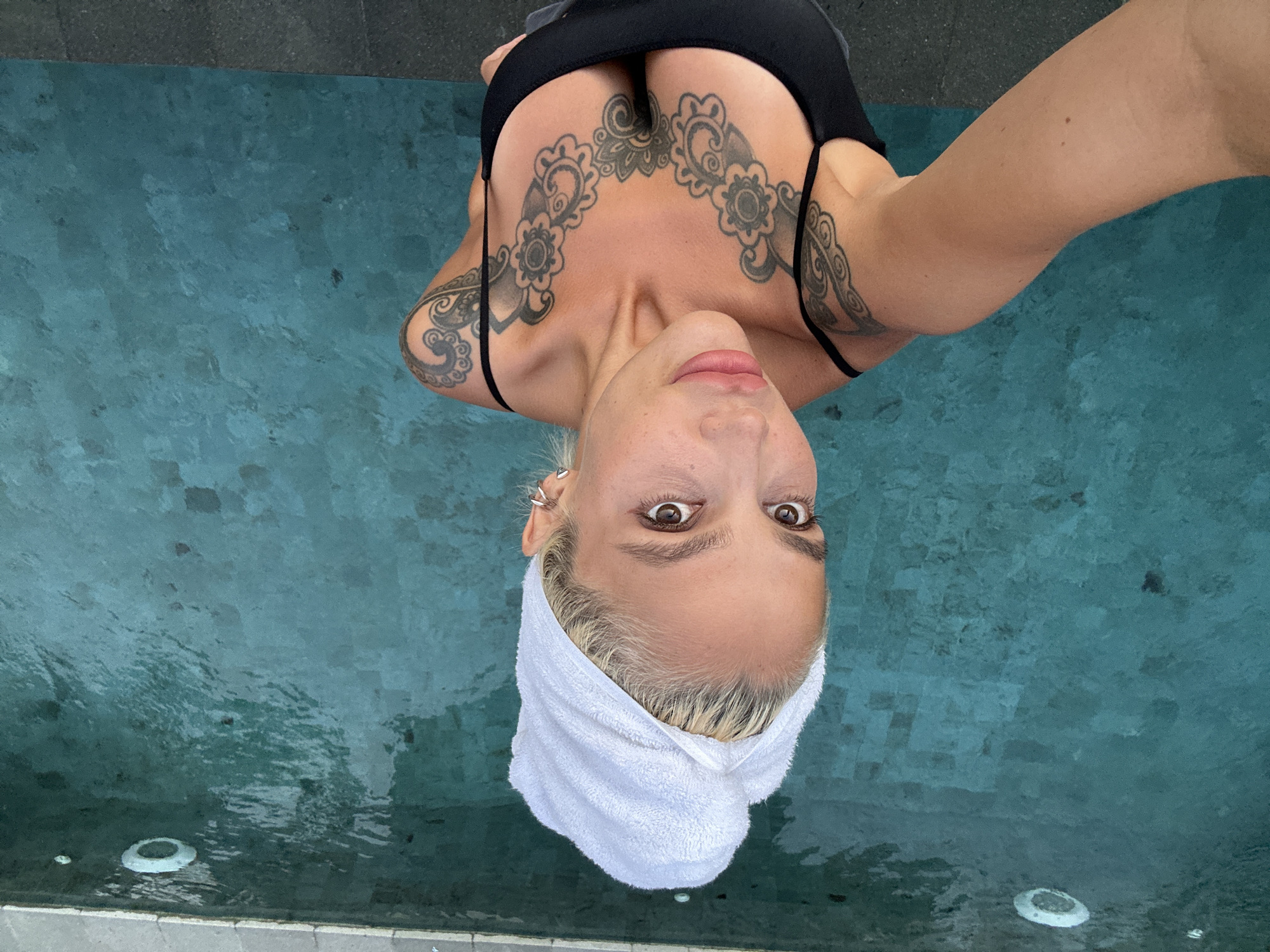 An overhead, medium-shot selfie captures a fair-skinned woman with blonde hair wrapped in a white towel, standing in a swimming pool. She is wearing a black bikini top, revealing extensive black tattoo work on her chest and shoulders, including intricate floral patterns. Her eyes are open and looking directly at the camera, and her lips are slightly parted. The pool water is a vibrant turquoise, with visible tile work at the bottom and a few small dark objects floating on the surface. The edge of the pool, composed of grey paver stones, is visible at the bottom of the frame. The scene appears to be outdoors, potentially at a resort or private residence in Kuta Selatan, Indonesia, given the style of the pool and architecture.