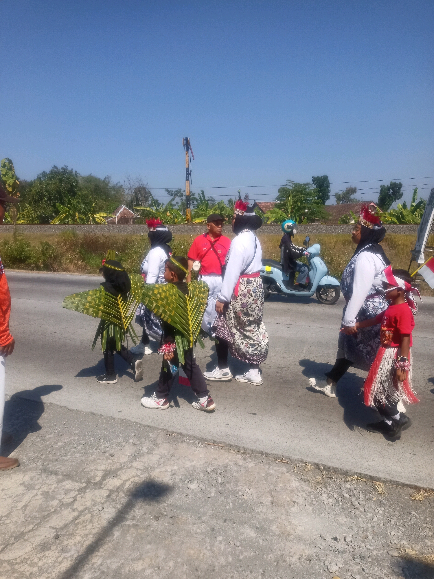 In Sumberlawang, Indonesia, on a clear, sunny day, a group of people are participating in a parade or procession. The scene unfolds on a paved road with a gravel shoulder, bordered by vegetation and trees. In the background, a railway line is visible, with a signal pole beside it.
The group consists of several individuals, including children dressed in festive attire. Two children in the foreground are wearing elaborate costumes made of green palm leaves, resembling wings or large fans attached to their backs and arms. They also have red and white decorative elements around their heads and waists.
Other participants include adults and an older child. One woman wears a white long-sleeved shirt, a patterned skirt, and a headscarf, adorned with a red and gold headband. Another adult, dressed in a red shirt and white pants, walks alongside. A younger child in a red shirt with a fringed skirt also stands out.
In the middle ground, a person is riding a light blue scooter, adding a modern element to the traditional procession. The overall atmosphere suggests a cultural event or celebration taking place outdoors. The sunlight casts distinct shadows on the road, indicating the time of day.