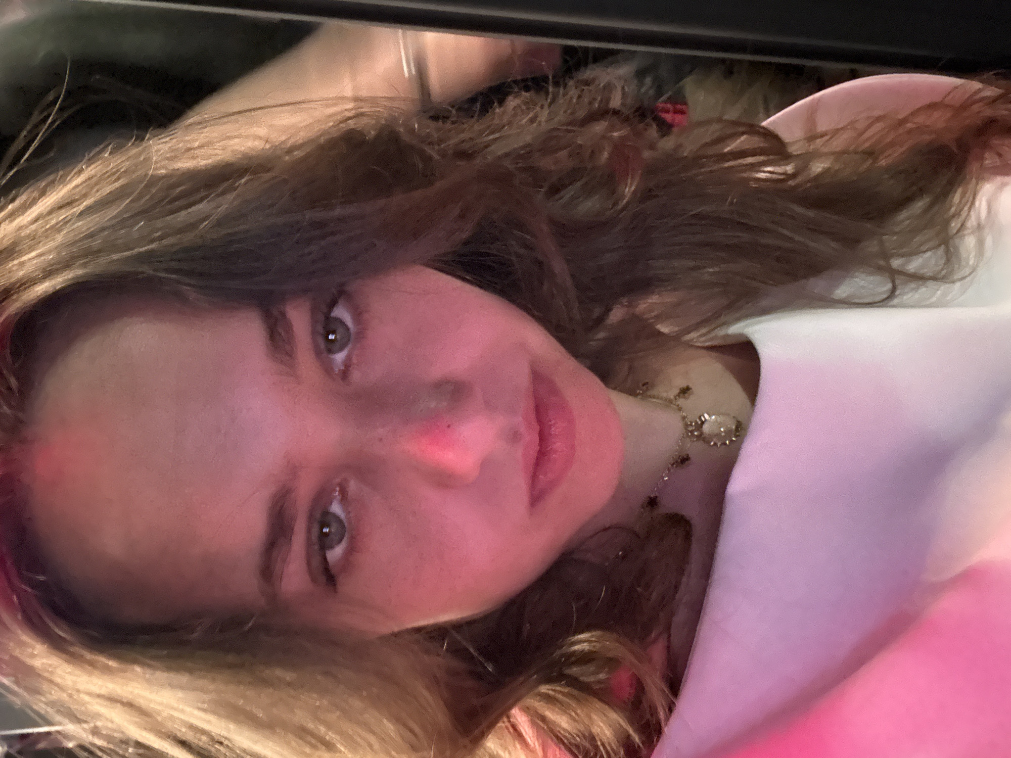 A young woman with long brown hair, wearing a white top and a necklace, is pictured in a close-up, angled shot. The background is dark, but there's a hint of light above her head. The lighting in the image appears artificial and possibly includes a pinkish hue illuminating her face. The location cannot be determined from the image.