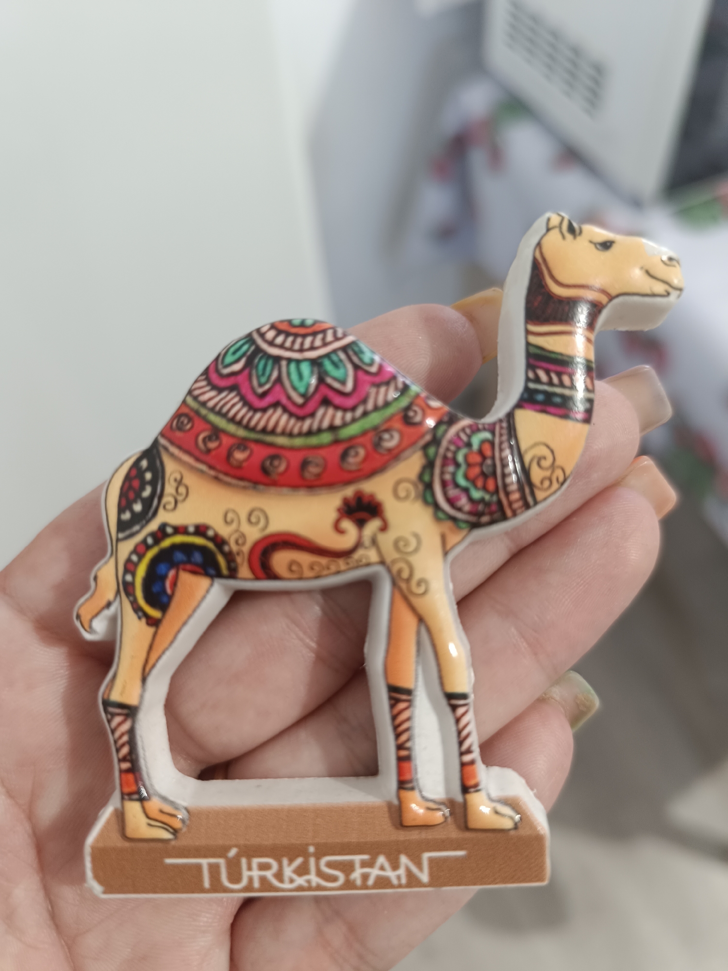 A person's hand is holding a camel figurine that says "TURKISTAN" on its base. The figurine is brightly colored with intricate patterns. The background is blurry and appears to be indoors. There is no indication of the city of Taraz, Kazakhstan in the image.