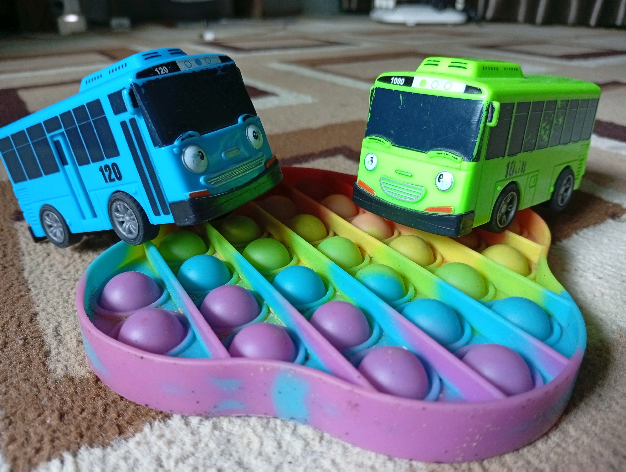 Two toy buses, one blue and one green, are placed on a tie-dye colored fidget toy with a guitar shape. The blue bus has the number "120" printed on its side and front. The green bus has the number "1000" printed on its front. The fidget toy has rows of colorful bubbles, predominantly pink, purple, blue, green, and yellow. The background consists of a patterned carpet. The scene is indoors. This image was taken in Kebondalem, Indonesia.