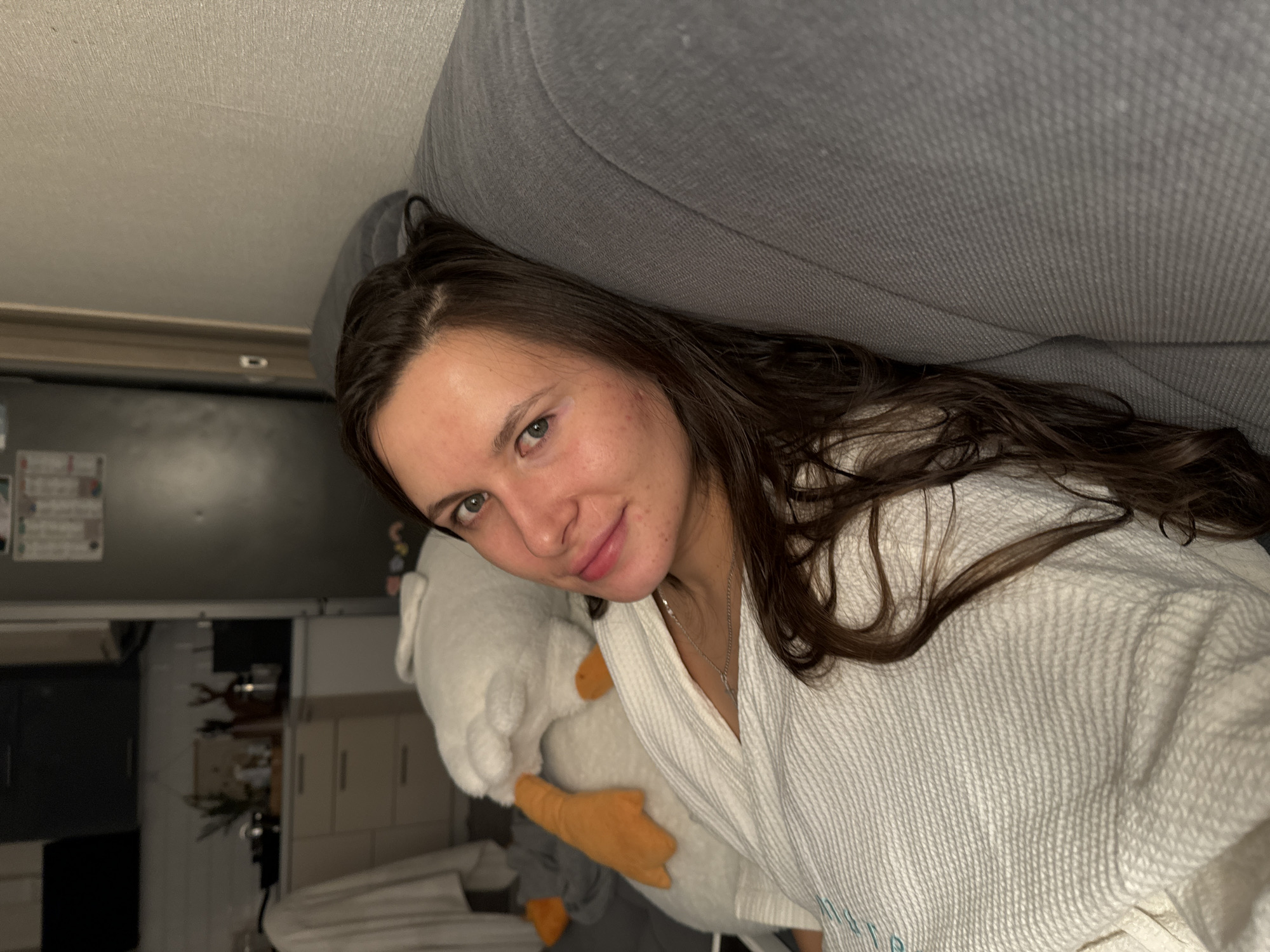 A young woman with long, dark brown hair and light eyes is pictured indoors, appearing to be lying down or reclining. She is looking directly at the camera with a gentle, relaxed smile. She wears a white, textured robe. Cradled in her arms is a large, white plush toy resembling a goose or duck, with noticeable orange feet and beak.
Behind her, a grey couch or headboard is visible, against which her hair is resting. The background on the left side of the image reveals what appears to be a domestic setting, possibly a kitchen or utility area. This area includes light-colored cabinetry, a dark surface, and various indistinct items, suggesting a lived-in space. The lighting is artificial and somewhat dim, indicating the photo was likely taken in the evening or at night. There is no discernible text visible in the image. The location context provided is Akulovo (Gp Kubinka), Russia.