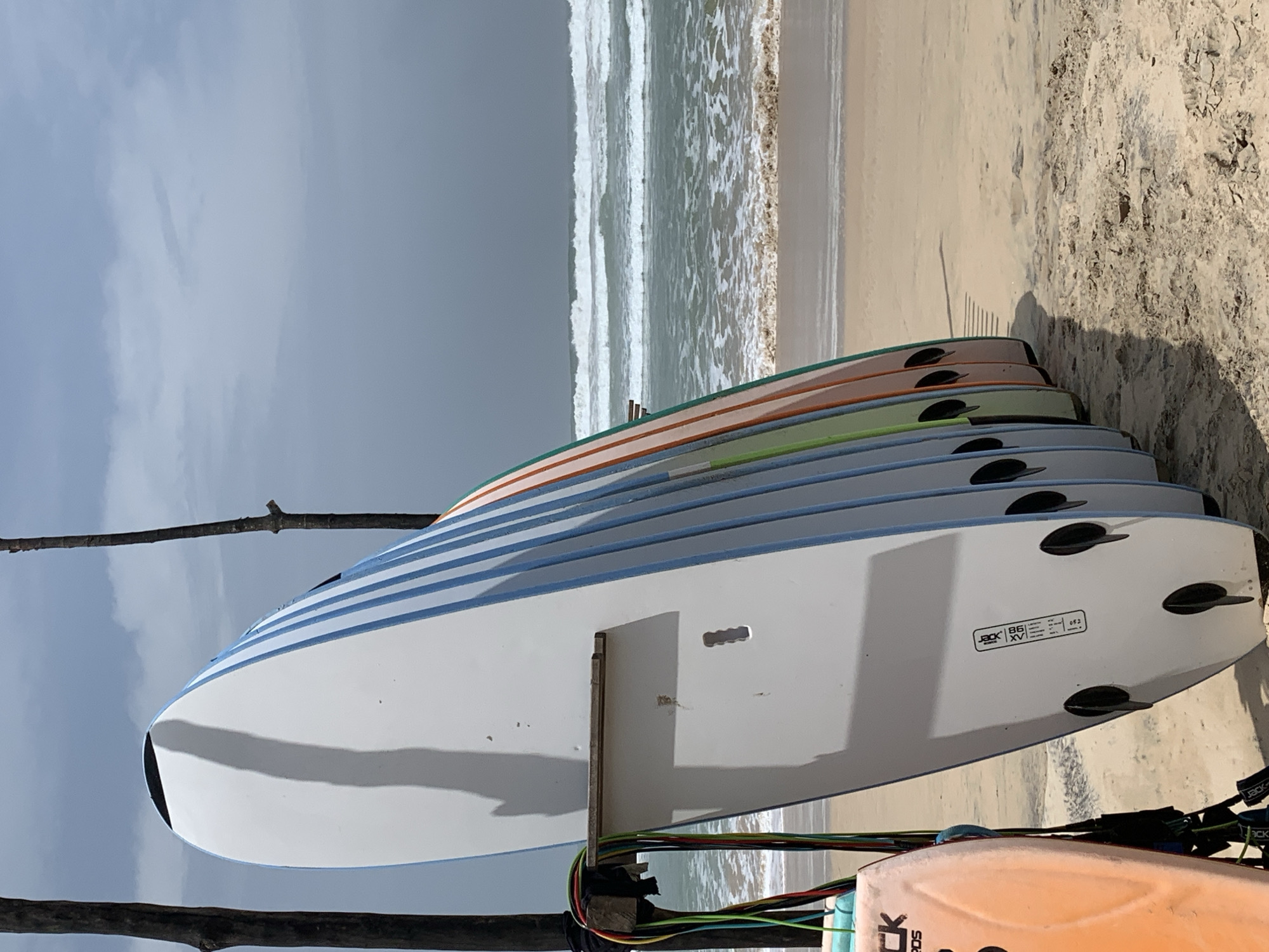 An array of surfboards stands upright on a sandy beach in Ahangama, Sri Lanka. The surfboards are lined up with their fins facing the viewer, some are white with blue stripes, others are in varying pastel colors like green, orange, and pink. Leashes in multiple colors hang from a wooden support structure attached to the foremost white surfboard. The ocean in the background exhibits moderate waves breaking on the shore under a cloudy sky. The sand is light brown, with tire tracks and scattered debris. The overall scene suggests a surf rental or school location.