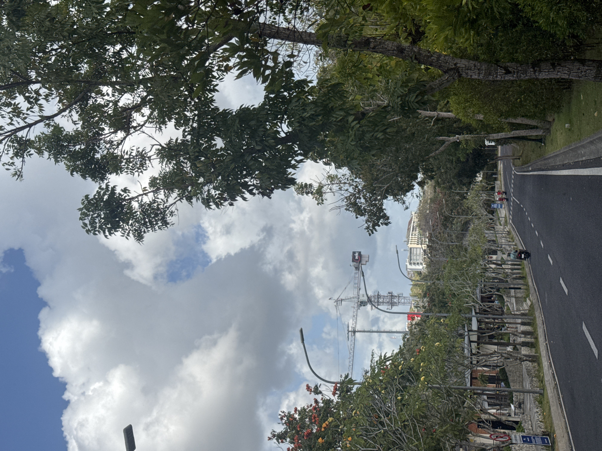 This is an outdoor, eye-level, medium shot photograph taken on a cloudy day in Kuta Selatan, Indonesia. The image captures a road with a slight curve, flanked by trees and some low-rise buildings on the left. On the right side of the road, dense greenery and trees are visible. In the distance, construction cranes are present, suggesting ongoing development. A single motorcycle with a rider is seen on the road, moving away from the camera. The sky is filled with large, white, puffy clouds.