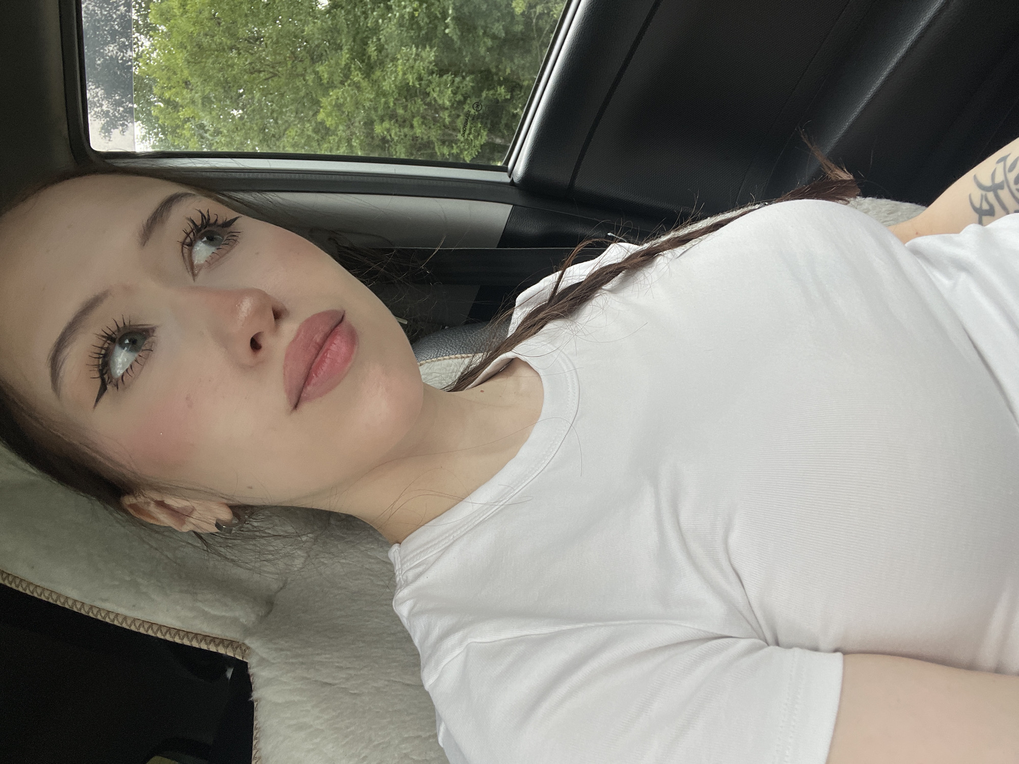 A young Caucasian woman with long brown hair in a braid, wearing a white t-shirt, is sitting in the passenger seat of a car. She is looking out the window to her right, with a slight smile. Her makeup includes winged eyeliner and pink blush. The car interior is visible, including a fluffy seat cover and the car door. Outside the window, trees are visible, suggesting a rural or suburban setting. The image appears to be taken during the daytime, under overcast skies.