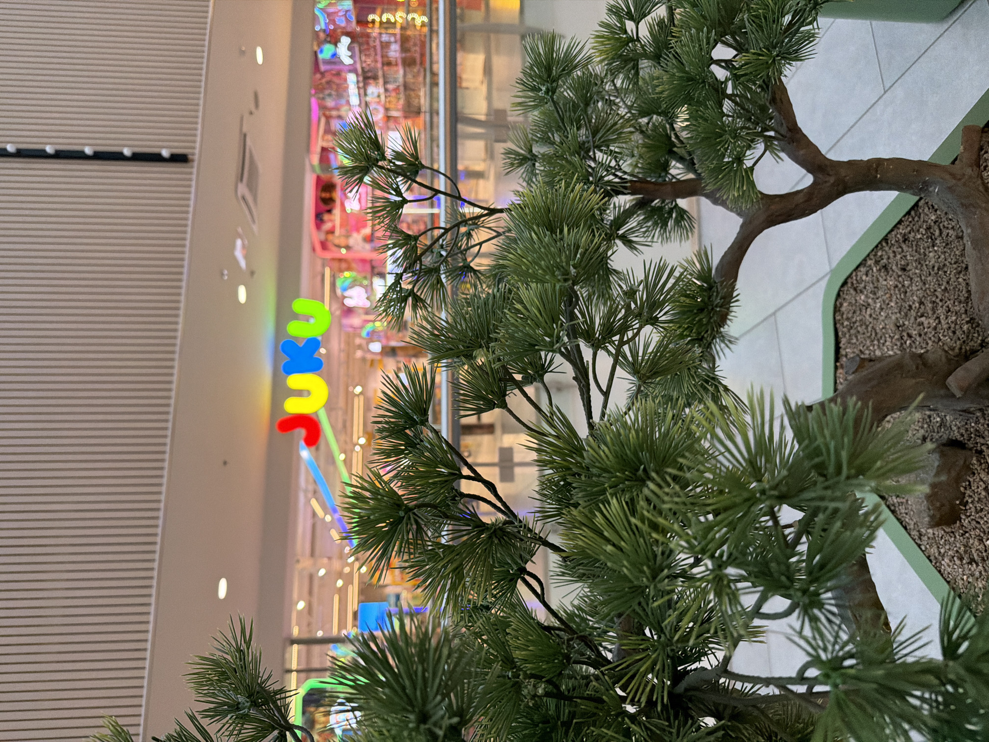 An artificial pine bonsai tree is in the foreground, its branches and needles obscuring a brightly lit storefront in the background. The storefront features a large, multi-colored sign that reads "JUJU" in bold, block letters. The background also shows shelves filled with various products, suggesting a retail environment, possibly a toy or novelty store. The lighting is warm and colorful, with streaks of blue and pink light visible. The flooring appears to be light-colored tiles. The overall scene is indoors, likely within a shopping mall or similar commercial establishment. There are no people visible in the image.