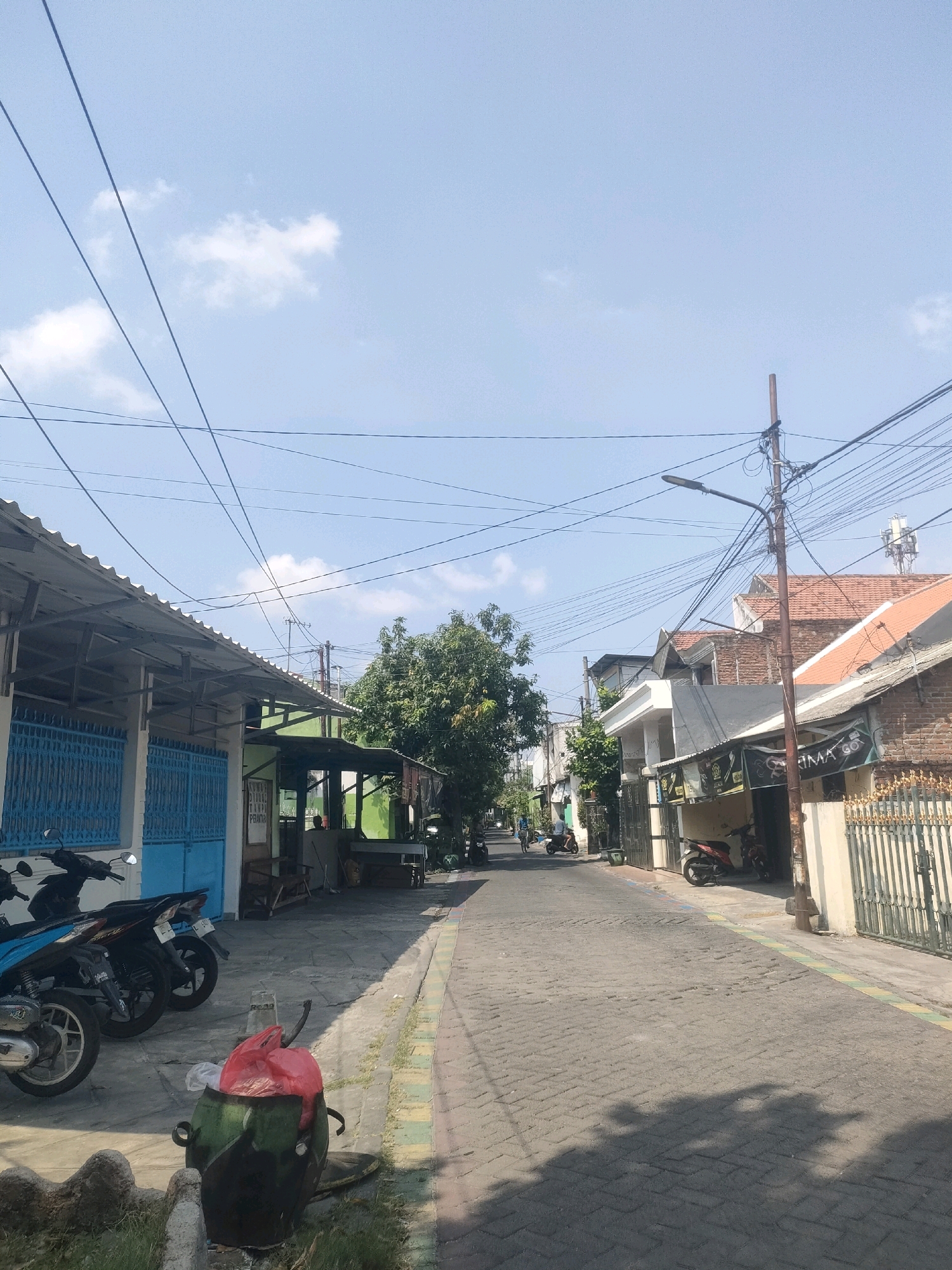 This is a vertical, eye-level, medium shot of a narrow street in Surabaya, Indonesia, on a sunny day with a clear blue sky and scattered clouds. The street is paved with grey interlocking bricks, and on both sides are residential buildings with various architectural styles and roof materials, including red tile and corrugated metal.
On the left side of the street, a building with a corrugated metal roof is visible. It has a light blue gate and several parked motorcycles in front. The building appears to be a shop or a business, with a sign that is partially obscured by a green awning.
On the right side of the street, a row of houses and businesses lines the road. Some buildings have red tile roofs, while others have darker colored roofs. There are decorative fences and gates in front of some of the properties. Several more parked motorcycles are visible along this side of the street.
A utility pole with numerous wires crisscrossing the sky stands in the middle ground on the right side of the street. A streetlamp is attached to the pole. In the background, more houses and trees are visible, extending down the length of the street.
In the foreground on the left, a green and red trash can is overflowing with garbage, with plastic bags visible. The street is mostly empty, with only a few indistinct figures and a couple of distant motorcycles suggesting some activity. The overall impression is of a typical residential street in a densely populated urban area.
