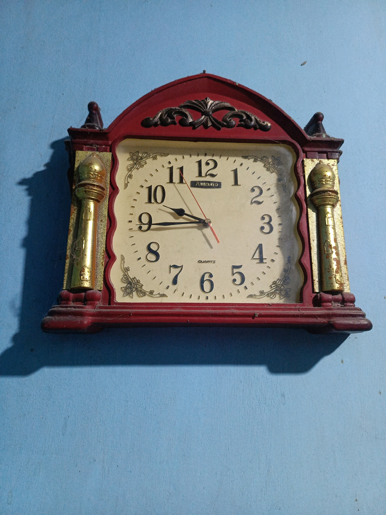 A vintage, rectangular wall clock with a red and gold ornate frame hangs on a light blue textured wall. The clock face is cream-colored with black numerals and decorative floral elements in the corners. The hour hand points to approximately 10:45, and the second hand is near the 5. The clock has a decorative finial at the top and two golden, fluted columns on either side of the face. The clock is slightly dusty, and a shadow of the clock is cast on the wall to its left. This clock is located inside a residential building in Kalang, Indonesia.