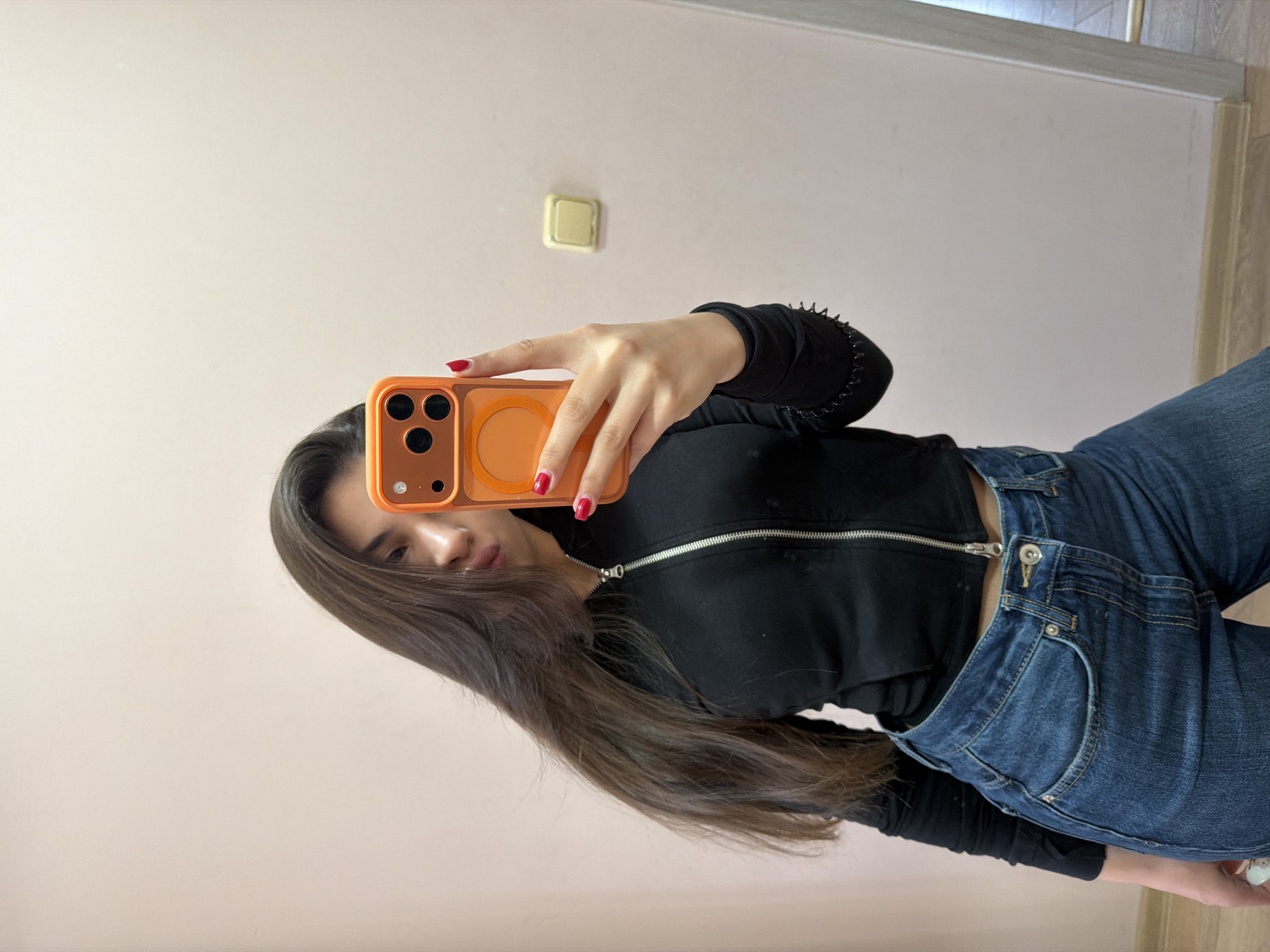 A young woman is depicted taking a mirror selfie in an indoor setting, likely in Rudartsi, Bulgaria. She holds an orange smartphone with a prominent camera array and a circular MagSafe-compatible ring in her right hand, partially obscuring her face, though her lower nose and lips are visible. She has long, dark brown hair that flows downwards. Her fingernails are painted with bright red polish. She is dressed in a black long-sleeved top with a silver zipper running down the front, paired with blue denim jeans.
The background features a plain, light-colored wall, possibly pale pink or beige, on which a gold-colored square light switch or power outlet is mounted towards the top left. The bottom of the image reveals a light-colored wooden floor separated from the wall by a white skirting board. The lighting appears to be natural, suggesting daytime. No discernible text, specific emotions, or particular weather conditions are evident in the image.