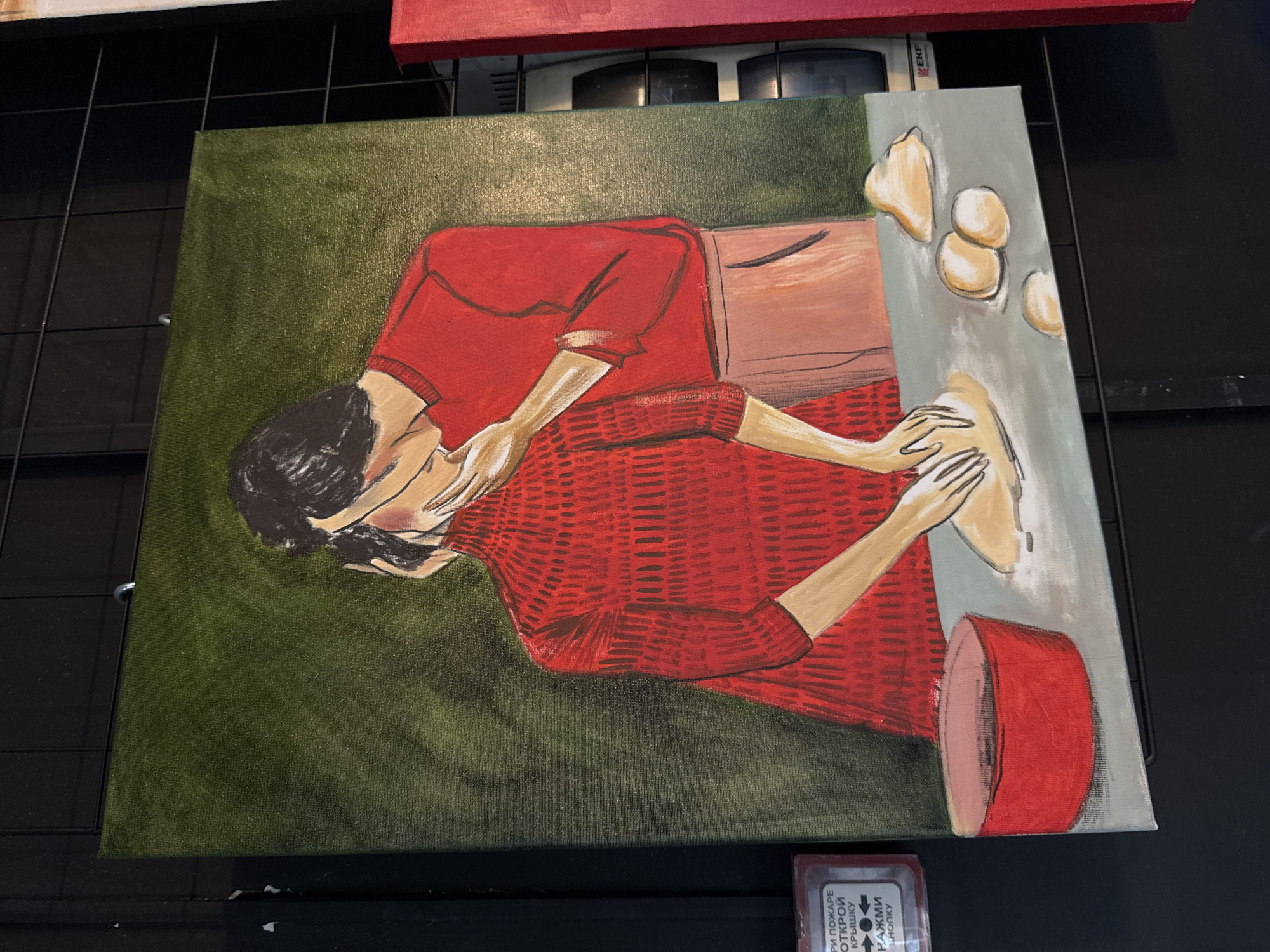 An illustrative painting on canvas, captured in Izhevsk, Russia, depicts a person engaged in food preparation. The subject, seen from the chest up, has dark hair and fair skin, wearing a textured red long-sleeved sweater and what appears to be a light brown apron or skirt. Their head is bowed, and their hands are actively kneading or shaping a large piece of light-colored dough on a light gray table or counter surface. Several smaller, irregularly shaped dough pieces are also present on the table. To the right, a red cylindrical container with a lighter interior rests on the surface.
The painting's background is a dark, brush-stroked green. The artistic style features bold outlines and distinct color blocking, with visible brushwork, particularly in the green background and the textured red sweater. The person's posture suggests concentration and focus on the task.
The canvas is displayed vertically, resting against a dark background, possibly a black wire rack or shelf. Hints of other red canvases are visible above. Below the painting, a small electronic device displays Cyrillic text: "ПРИ ПОЖАРЕ ОТКРОЙ КРЫШКУ НАЖМИ КНОПКУ," which translates to "IN CASE OF FIRE OPEN LID PRESS BUTTON," indicating an emergency or fire alarm device.