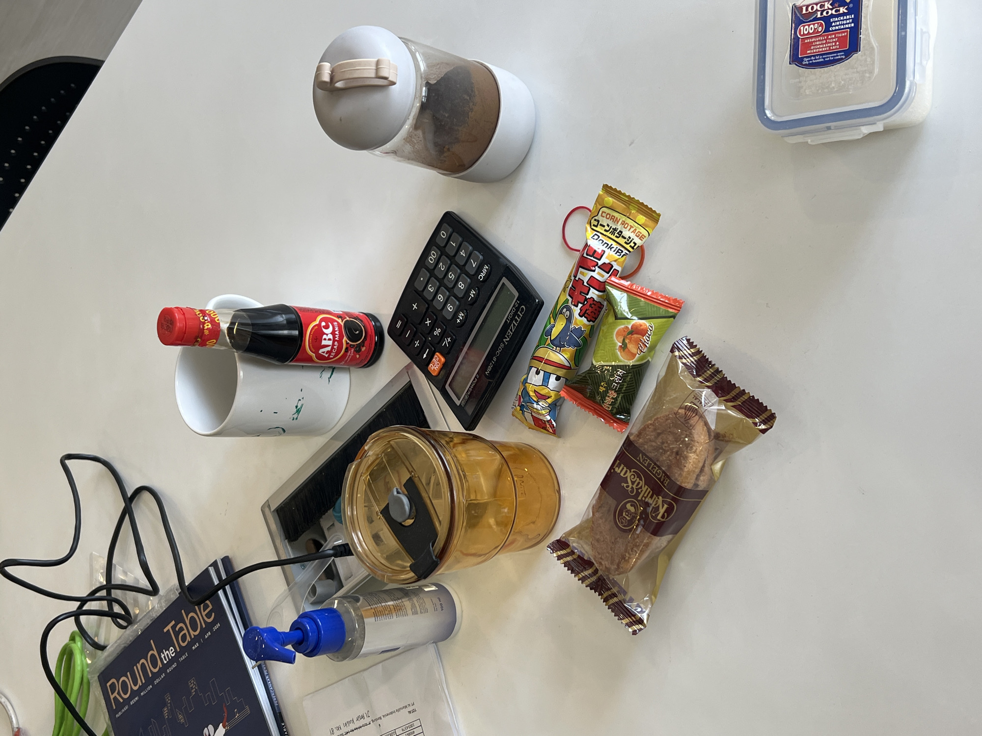 The image depicts a white table with various objects, including a book titled "Round the Table," a blue spray bottle, a yellow tumbler, a calculator, a bottle of ABC Kecap Manis, a mug, several snack wrappers, and a white food storage container. The environment appears to be an indoor setting, possibly an office or home. There are no people or animals visible in the scene. The lighting is bright and even, suggesting it might be daytime.