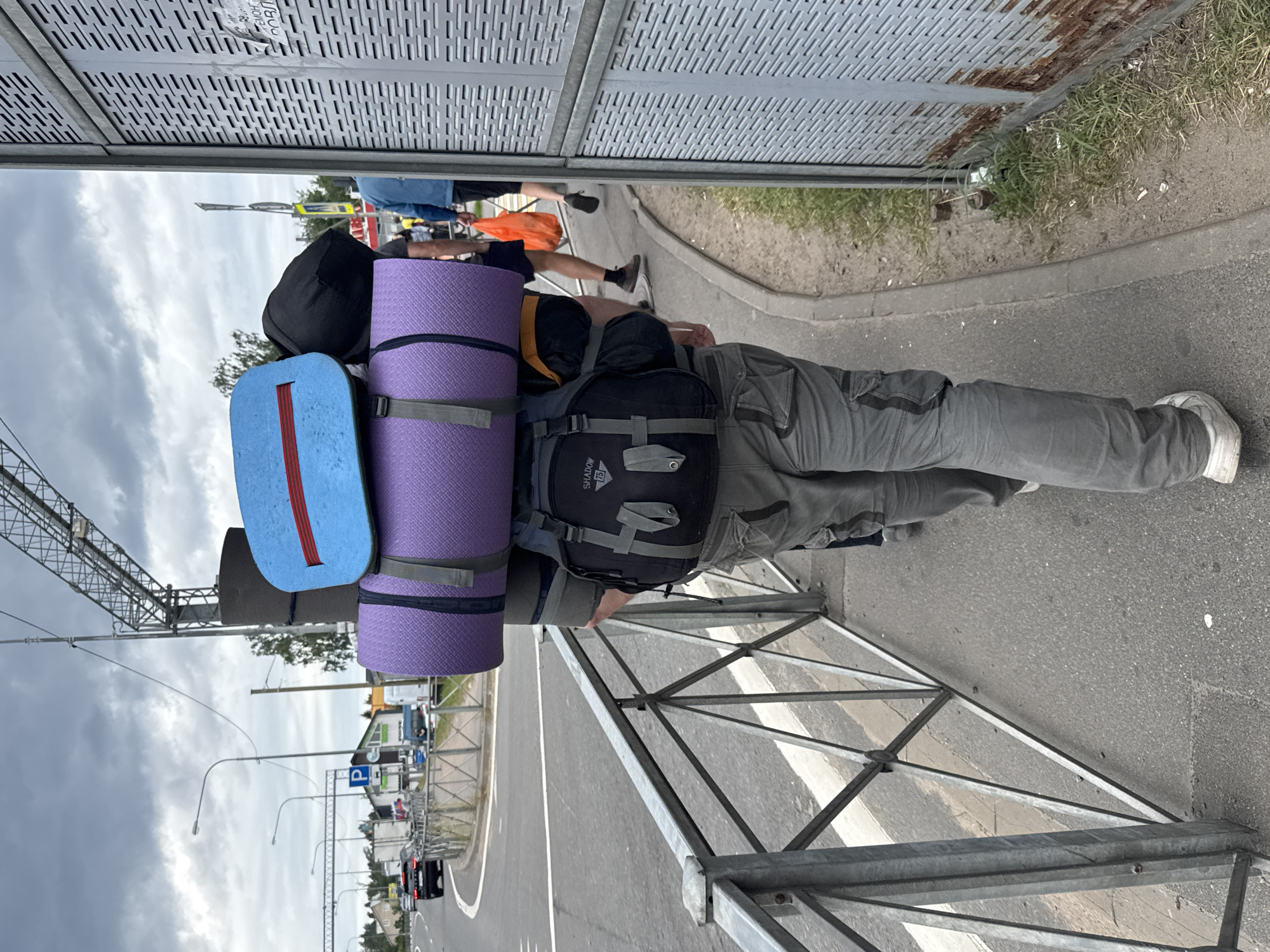 A person, viewed from behind, walks away from the camera on a sidewalk next to a road. The person is heavily laden with camping gear, including a large black backpack, a purple sleeping mat strapped to it, and a blue rectangular object on top of that. The person is wearing grey cargo pants and white sneakers. The environment appears to be on the outskirts of Saint Petersburg, Russia, with a road, traffic, streetlights, and buildings visible in the background. A metal railing runs along the edge of the sidewalk, separating it from the road. A large, grey, perforated metal structure, possibly a sound barrier or fence, is on the right side of the frame. The sky is overcast with clouds. Other pedestrians are visible further down the sidewalk, one carrying a bright orange bag.