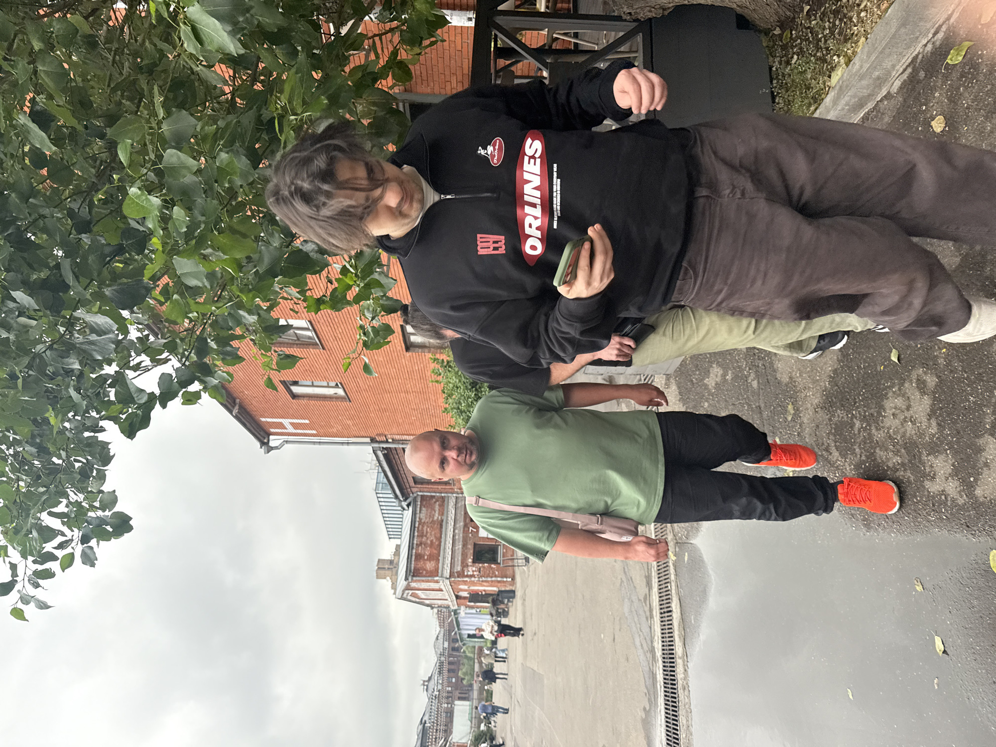 Two men walk on a wet paved area in an urban setting, possibly an industrial or creative district. The man in the foreground, on the right, is looking at his phone. He wears a black zippered sweatshirt with red and white graphics, including the word "ORLINES" and a year "1997", and dark brown baggy pants. The man on the left, facing the camera, is bald and wears a green t-shirt, black pants, and bright orange sneakers. He carries a light pink bag over his shoulder. In the background, several other people are walking in the distance. The architecture includes brick buildings, one with a white letter "H" visible on its facade. Trees with green foliage are present. The ground is wet, indicating recent rain. The sky is overcast.