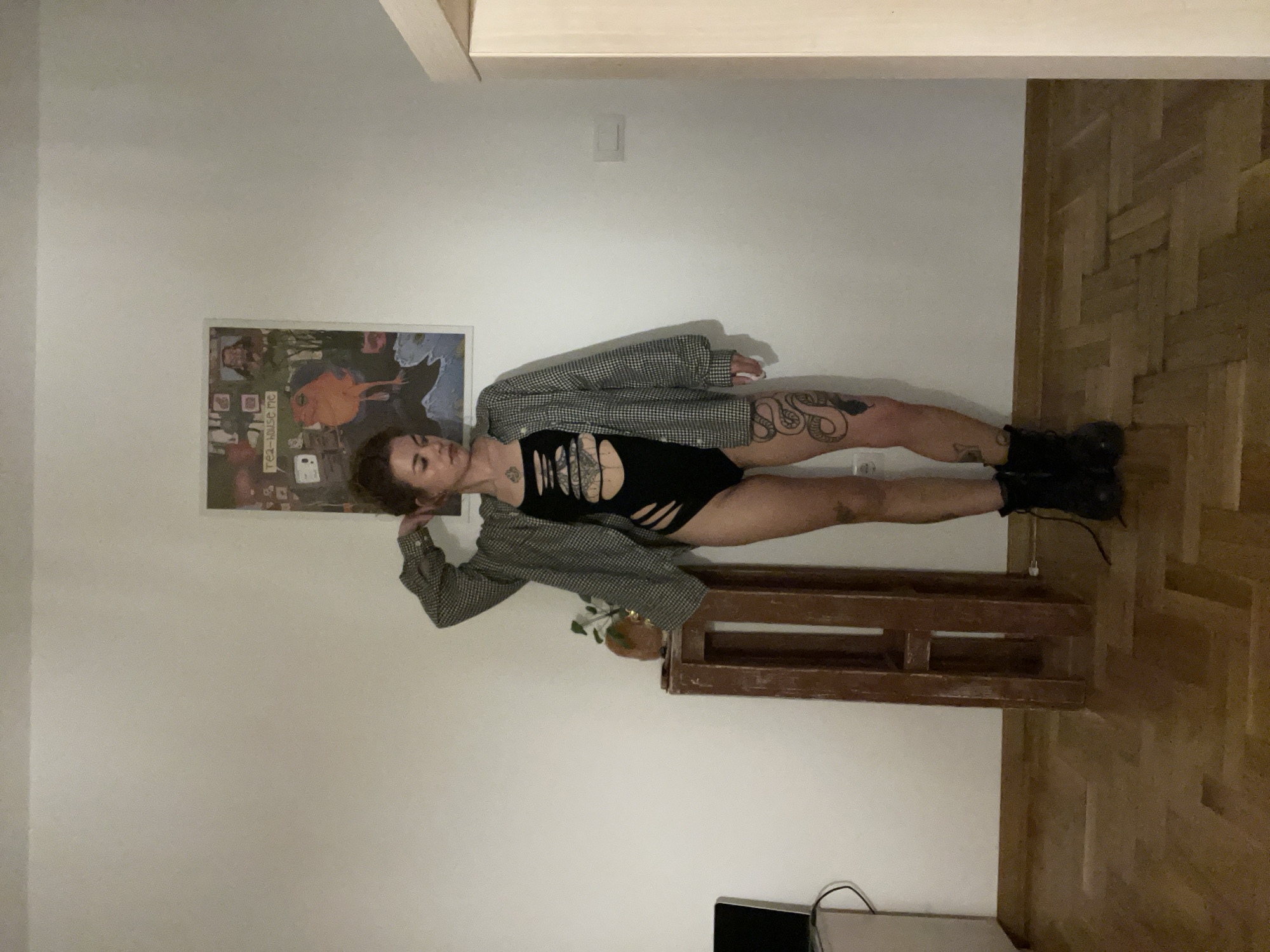 In an indoor full-body shot, a young woman poses against a plain white wall in Boreti, Montenegro. She stands upright, her dark hair slightly messy, gazing towards the viewer with a neutral expression. Her physique is adorned with numerous tattoos, notably a large snake design winding up her right thigh and hip, and abstract patterns on her left arm and calf.
She wears a distinctive black, cut-out bodysuit or swimsuit, revealing segments of her torso. A grey and white checkered blazer, possibly houndstooth, is draped over her shoulders and arms. Her feet are clad in black, chunky boots. Her pose involves a slight bend in her left knee, while her right foot rests on a dark brown wooden crate or pallet-like structure. A small potted plant with green leaves sits atop this wooden prop.
The setting reveals a light-colored parquet floor with a geometric pattern. Above, a wooden beam or ceiling structure is visible. On the wall to the left of the woman, a framed poster or artwork hangs, featuring an array of illustrations including figures, abstract designs, and a prominent orange character. Faint visible text on the poster includes "Tea-house me". A light switch is also visible on the white wall. The lighting appears to be soft and even, indicative of an indoor environment, possibly illuminated by artificial light.