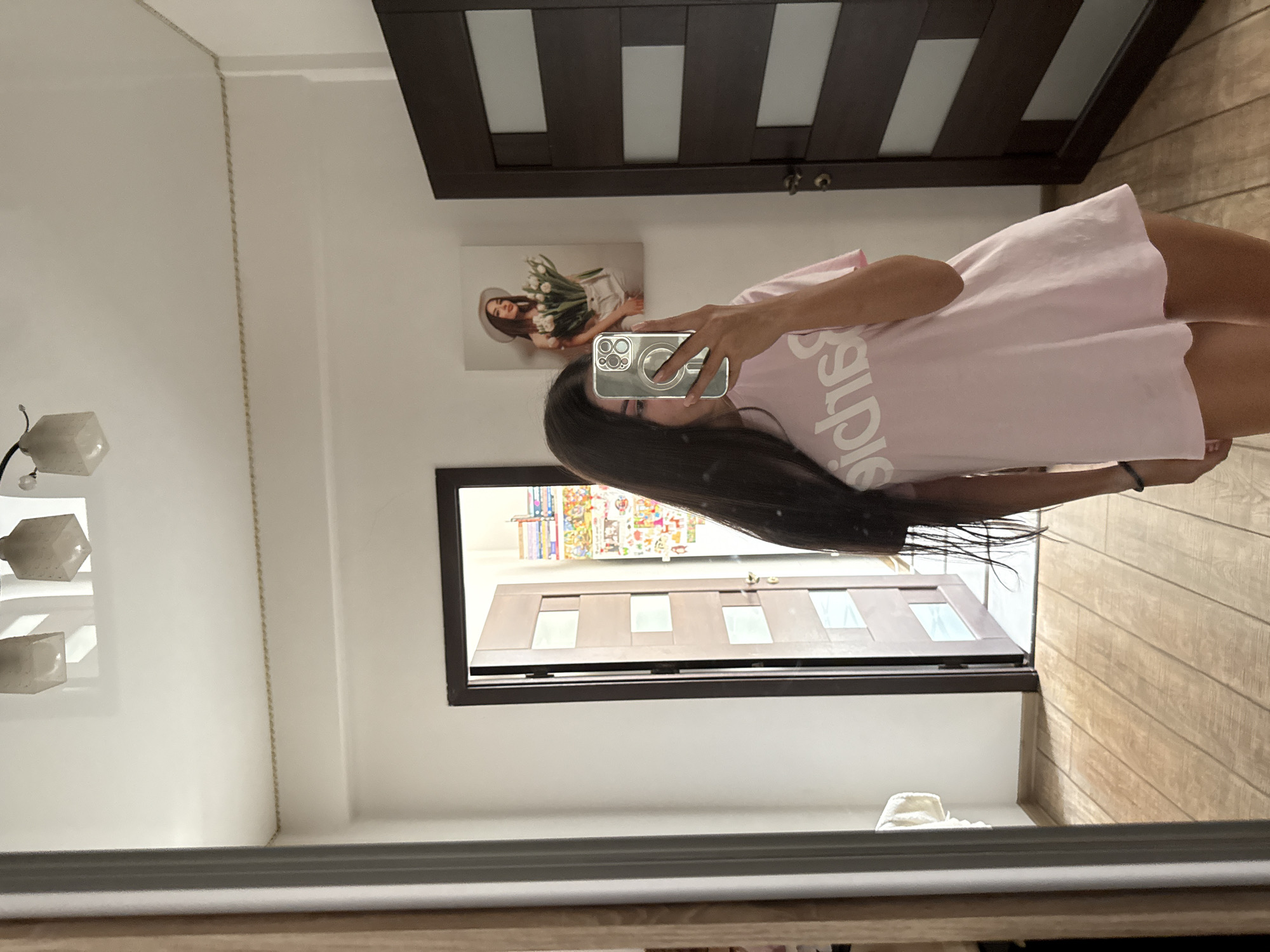 A young woman with long, dark hair is taking a mirror selfie in what appears to be a hallway or bedroom. She is wearing a light pink t-shirt dress with white text that reads "Barbie" across the chest. She is holding a clear phone case in her right hand. To her left, a large mirror reflects the scene. To her right, a dark wooden door with frosted glass panels is visible. Further down the hallway, another door is ajar, revealing a colorful mural or poster on the wall behind it. Above the woman's head, three square light fixtures are mounted on the ceiling. The floor is made of light-colored wooden planks. The room is well-lit, with natural light entering from an unseen source. The overall impression is a casual, domestic setting.
I am unable to determine the location of this image within the city of Chișinău, Moldova, as there are no specific landmarks or identifying features present.