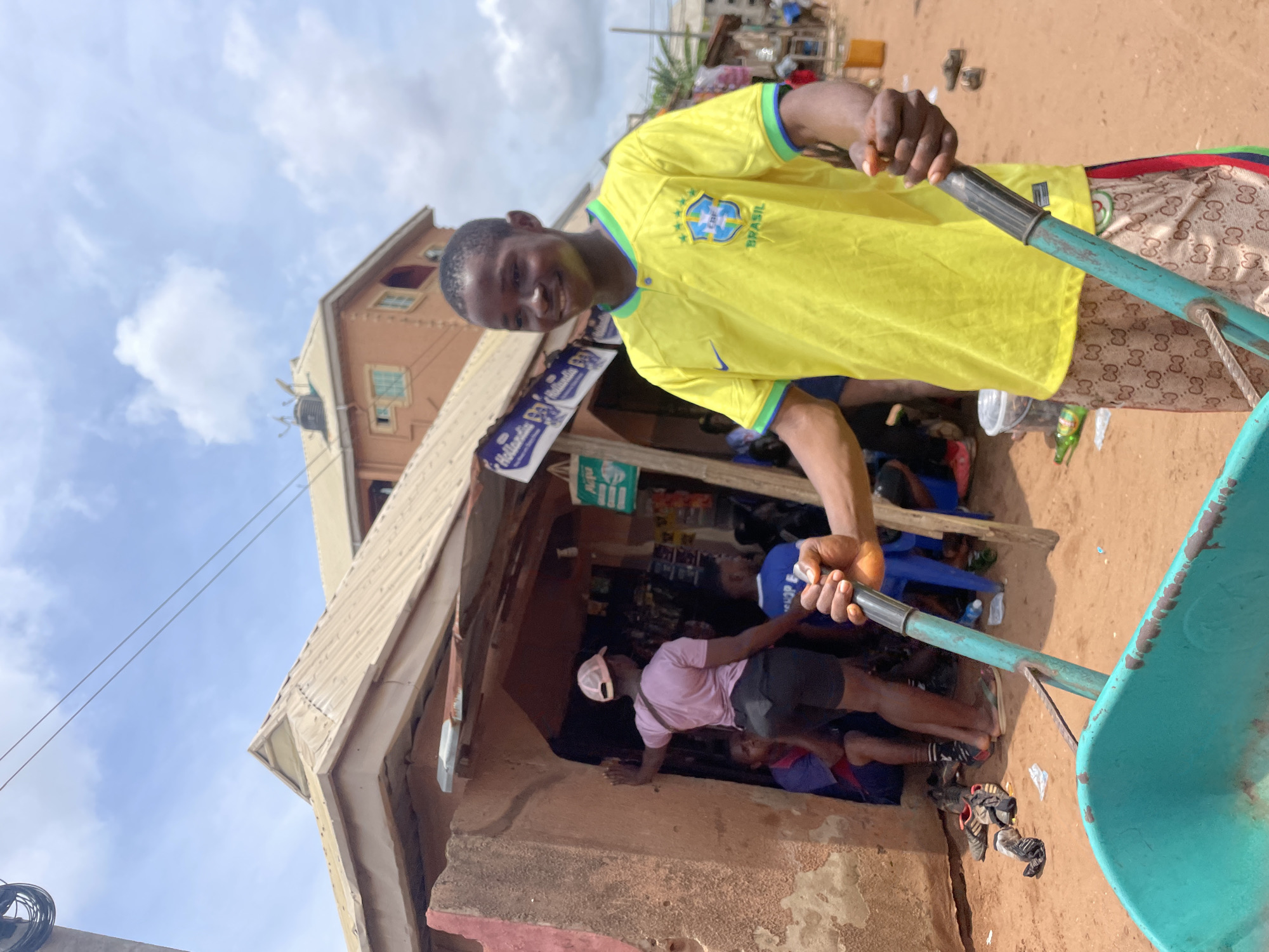 A man in a yellow Brazil soccer jersey is standing near a rough concrete building in Nkpor, Nigeria. He is holding the handles of a blue wheelbarrow, partially obscuring the view into the building's entrance. Inside, two other people are visible, one wearing a pink shirt. The building appears to be a small shop, with advertising signs visible. The surrounding environment consists of a sandy, dusty ground and other buildings in the background. The sky is partly cloudy.