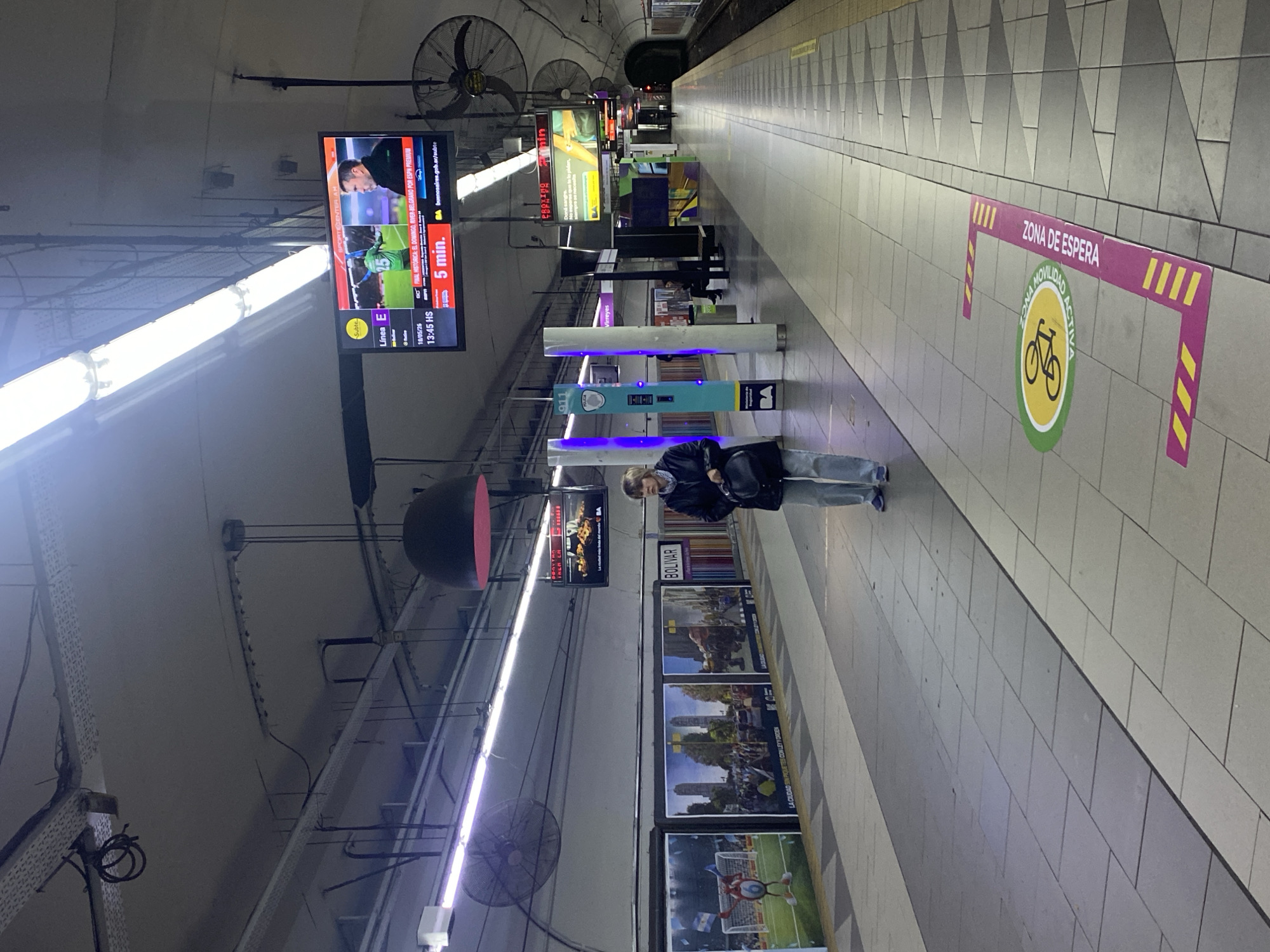 Here's a detailed description of the image:
The image depicts a subway station platform in Buenos Aires, Argentina. The platform is well-lit and tiled. Along the platform's edge, there are advertisements and signage, including one indicating "BOLIVAR." A screen displays a soccer game, the time (13:45 HS), the date (18/05/26), and an indication of a 5-minute wait. Safety features like a "ZONA DE ESPERA" (waiting area) sign with a bicycle symbol are visible.
A person is standing on the platform. There are lights, fans, and overhead cables visible on the ceiling. Emergency telephone and information pillars are also present.