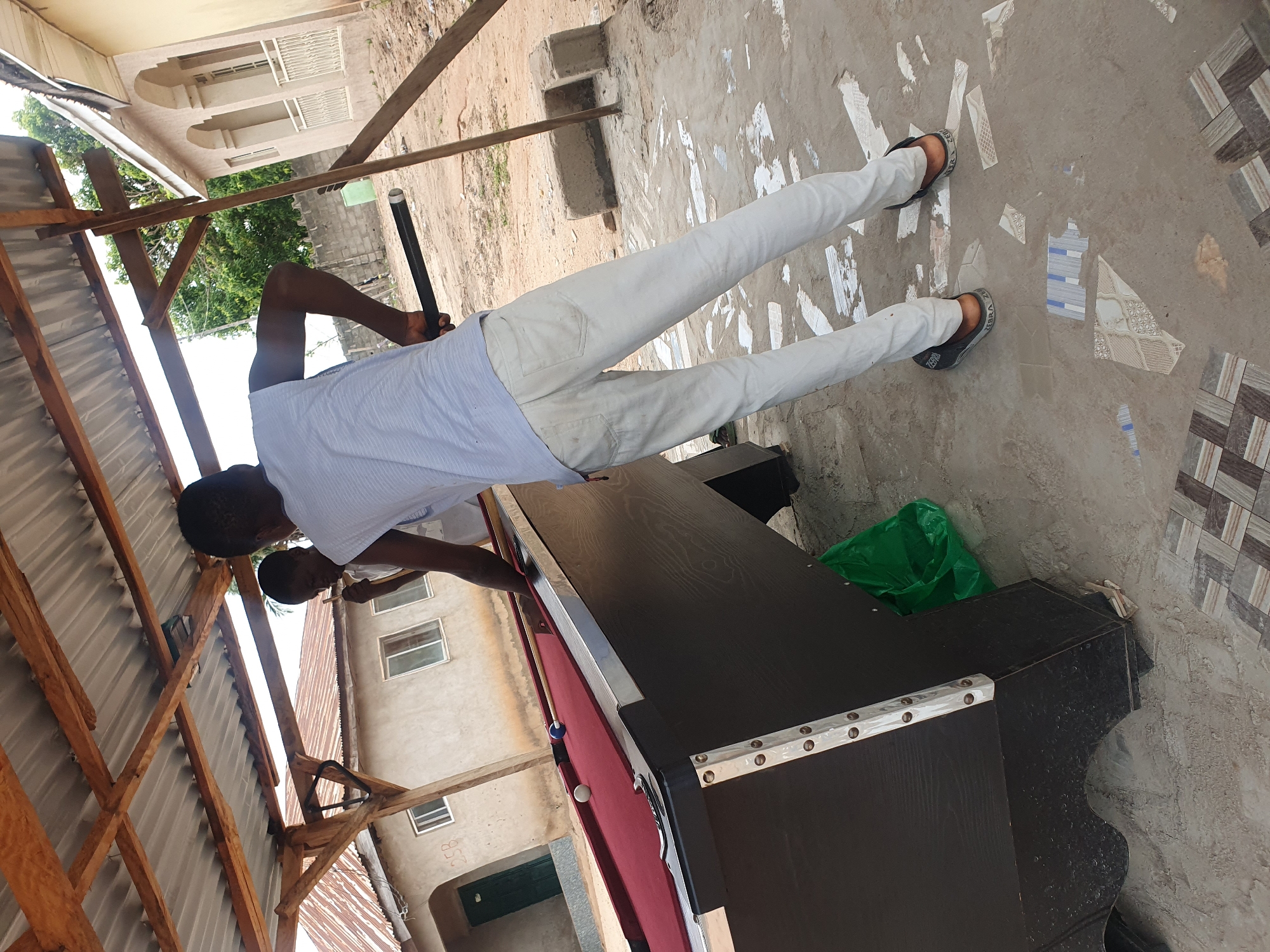In Lokoja, Nigeria, a man dressed in white trousers and a blue and white striped shirt stands on a concrete ground with scattered tile fragments. He holds a pool cue in his right hand, positioned over a burgundy pool table. The pool table has a black frame and appears to be outdoors, partially covered by a wooden structure with a corrugated metal roof. In the background, a light-colored building with multiple windows and balconies is visible. Another person, wearing a light-colored shirt, is partially visible on the left, also near the pool table. A green plastic bag is on the ground next to the pool table.