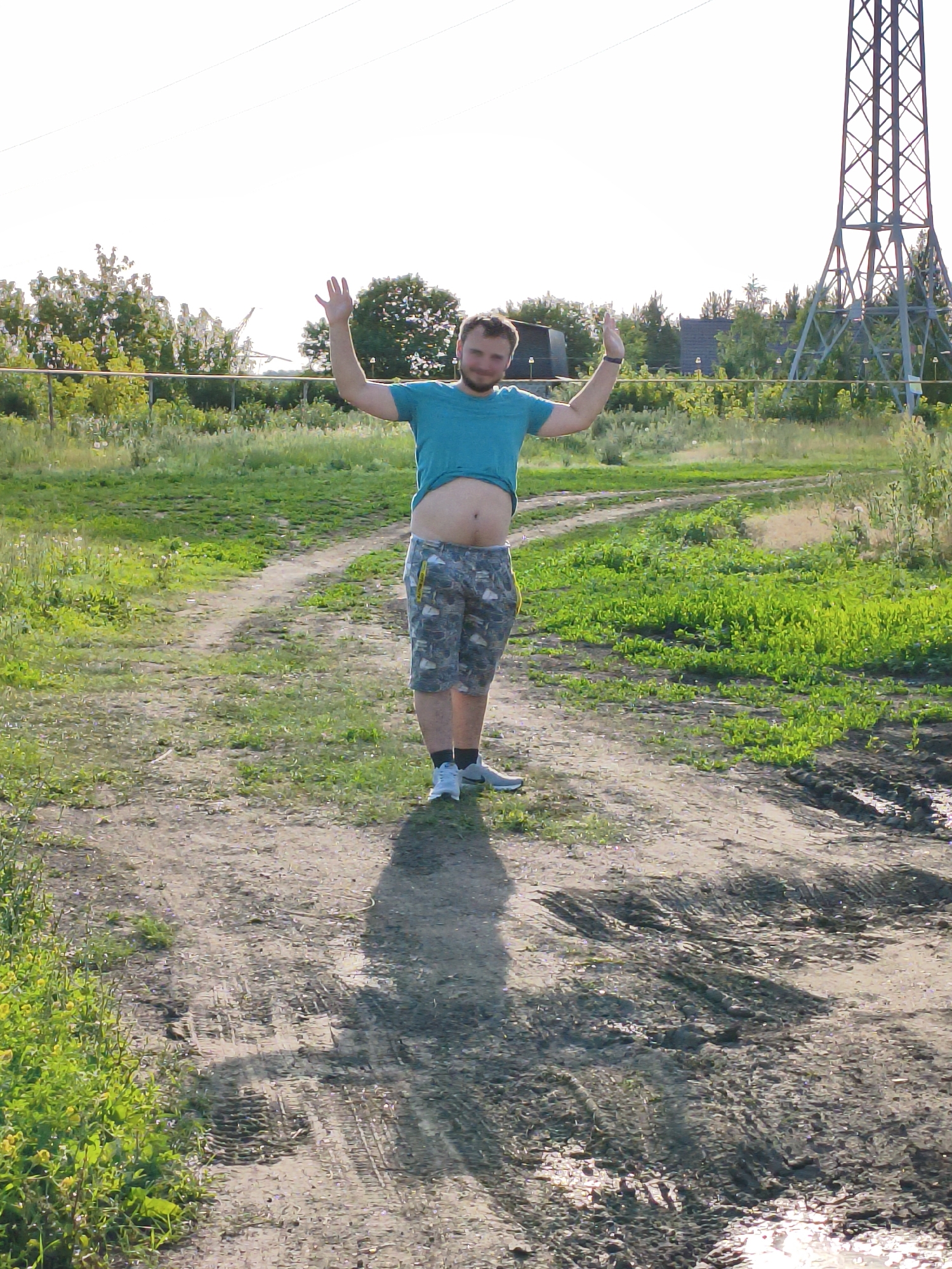 In Orenburg, Russia, on a dirt road with tire tracks and puddles of water, a man with short brown hair and a beard stands in the middle of the road. He is wearing a turquoise t-shirt that is lifted to expose his abdomen and camouflage-patterned shorts. The man has his arms raised and hands open. To the right of the road, there is a large metal pylon. The background consists of green grass, trees, and a bright sky.