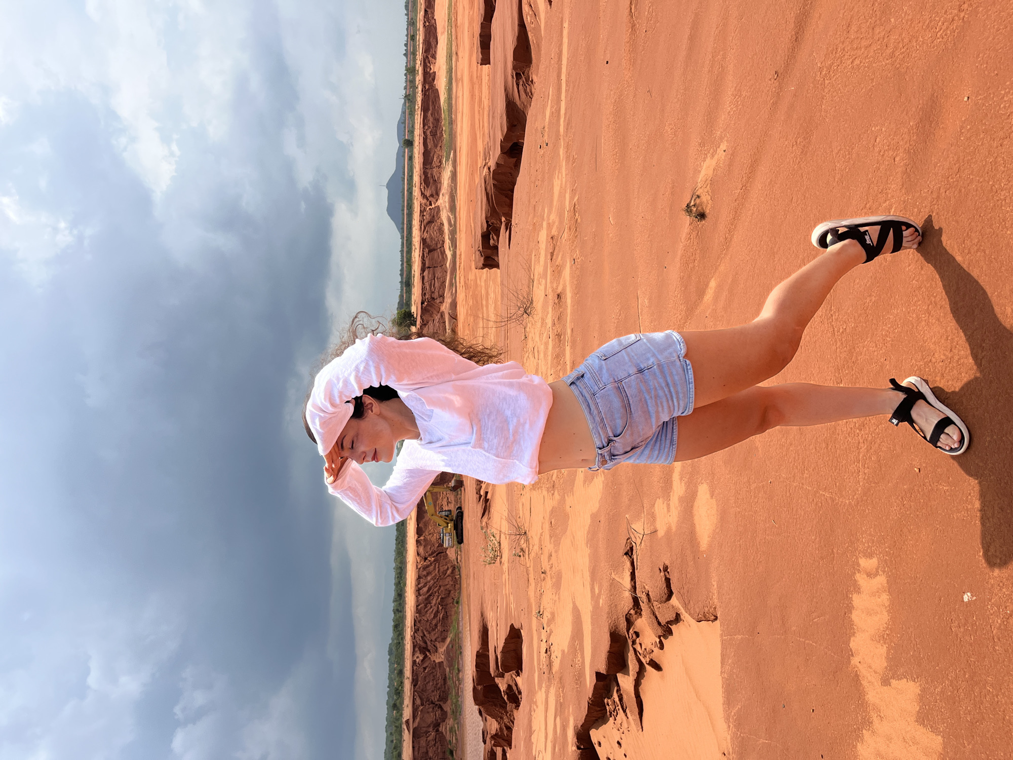 A young woman stands in a sandy, eroded landscape, her hands shielding her eyes as she looks out towards the horizon. She is wearing a long-sleeved white shirt and light blue denim shorts, with black and white sandals on her feet. The environment is characterized by reddish-orange sand, with visible erosion patterns and sparse vegetation. In the background, a yellow excavator is parked near a reddish cliff, and beyond that, a dark, cloudy sky looms over a distant mountain and trees. This scene is typical of the Red Sand Dunes in Mui Ne, Vietnam, a popular tourist destination known for its unique geological formations and photographic opportunities.