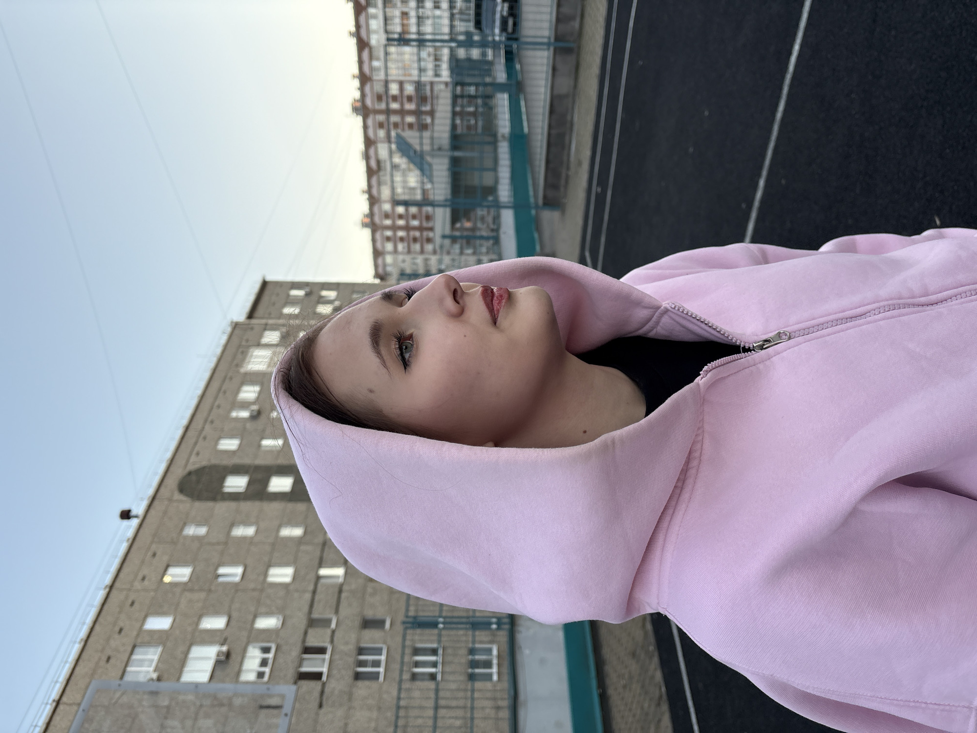 A young woman wearing a light pink hooded sweatshirt is looking up and to her right, with a neutral expression. The woman's skin is fair, and her eyes are light-colored. The buildings of Izhevsk, Russia, surround the subject. Behind her is a basketball court, followed by a red and white brick apartment building. To the side is a Soviet-era, tall, gray apartment building. The sky is overcast, and the light is diffuse. The photo is taken from a low angle, causing the background buildings to appear large and imposing.