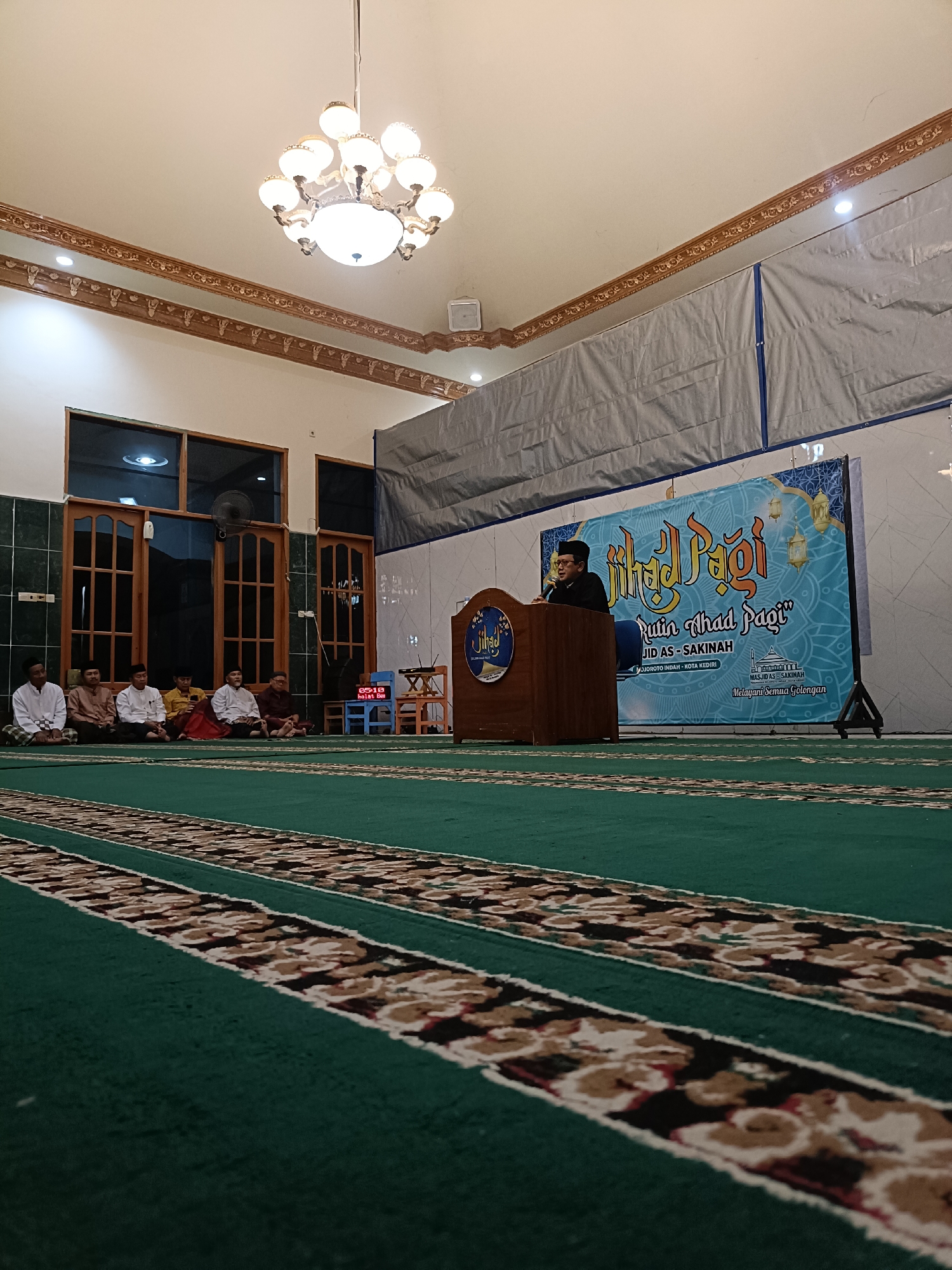 The image depicts an indoor event taking place in a mosque, likely the Masjid AS-SAKINAH in Kediri City, Indonesia, as indicated by a banner in the background. The scene is set on a green carpet with intricate patterned borders. In the foreground, the carpet is shown from a low angle, emphasizing its texture and design.
Further back, a wooden podium stands on the carpet, with a speaker wearing glasses and a black cap addressing an audience. Behind the podium, a large blue banner with the text "Jihad Pagi" and "Rutin Ahad Pagi" is displayed, along with imagery of lanterns and geometric patterns, typical of Islamic cultural events. The banner also includes text identifying the mosque and its location.
To the left of the podium, a group of men are seated on the floor, dressed in traditional Islamic attire. Some are looking towards the speaker, while others appear to be engaged in conversation or prayer. Behind them, large wooden-framed windows and doors suggest the architectural style of the building. The ceiling features a decorative cornice and a prominent chandelier. The overall atmosphere suggests a religious gathering or lecture.