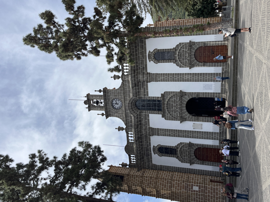 The image shows the Basilica de Nuestra Señora de la Candelaria in Teror, Gran Canaria, Spain. The basilica is constructed of grey stone with white accents. It features two side doors and a large entrance, all with wooden doors. There is a clock face on the facade above the entrance and a bell tower. There are two trees on the left side of the image. There are several people standing in front of the building, some looking at their cell phones and others interacting with each other. The ground is made of grey cobblestones.