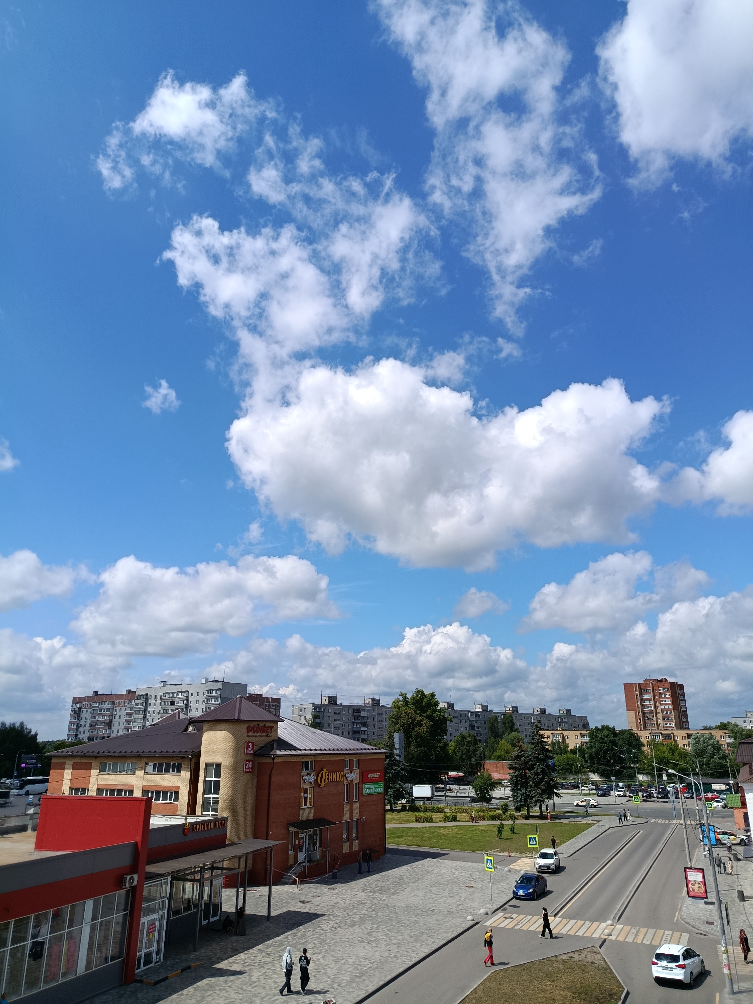 The image captures a street scene in Orekhovo-Zuyevo, Russia, under a bright, blue sky dotted with fluffy white clouds. In the foreground, a brick building with a brown roof features signage that includes "ОптОпт" and "Ленико". Adjacent to it, a building with a red facade is visible. A paved area with cobblestones and a road with markings are present. Several cars, including a white sedan and a blue sedan, are on the road. Pedestrians are seen walking on the sidewalks and crossing the street. In the background, multi-story residential buildings and trees form the urban landscape. The overall impression is a typical daytime scene in a Russian city.