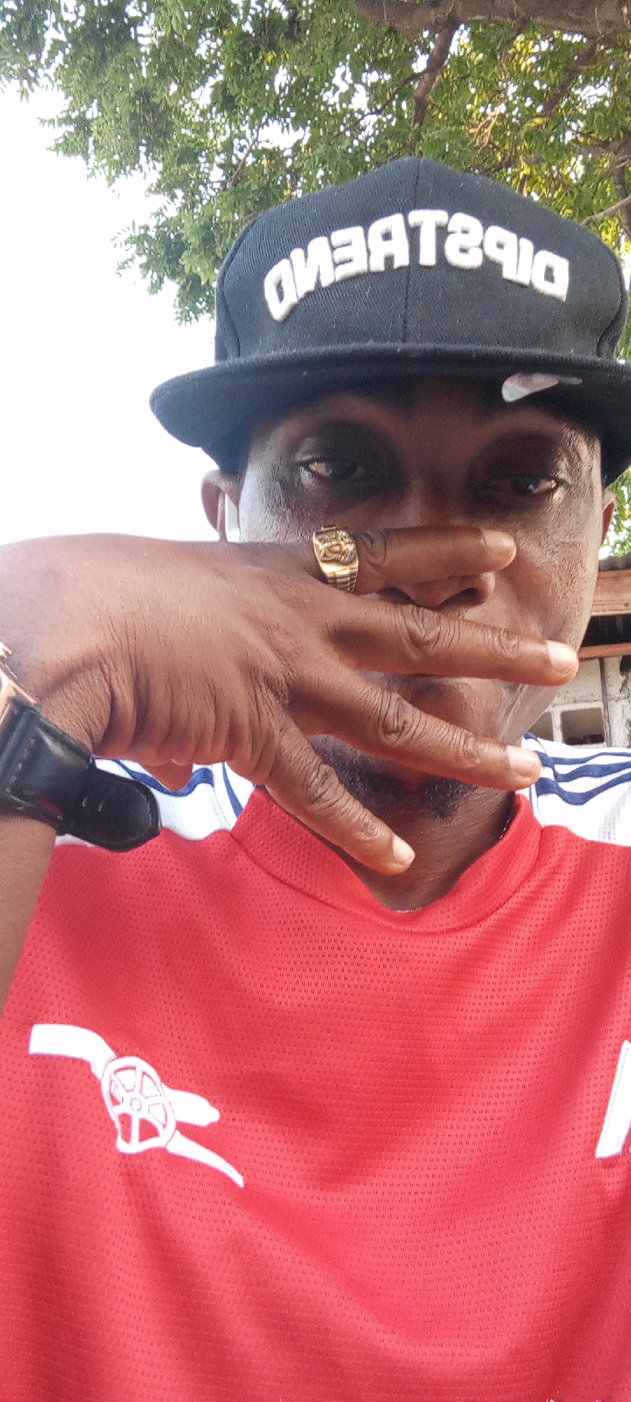 A man is posing in a close-up selfie. He is wearing a black baseball cap with the word "DISTRACTIONS" stitched in white, a red Arsenal soccer jersey with white accents, and a large, gold ring on his left ring finger. A black wristwatch is visible on his left wrist. He has his left hand raised, partially covering his face from the nose down. The background shows partial views of trees and buildings under a bright, overcast sky. The photo was most likely taken during the day in Zango-ndaji, Nigeria.
