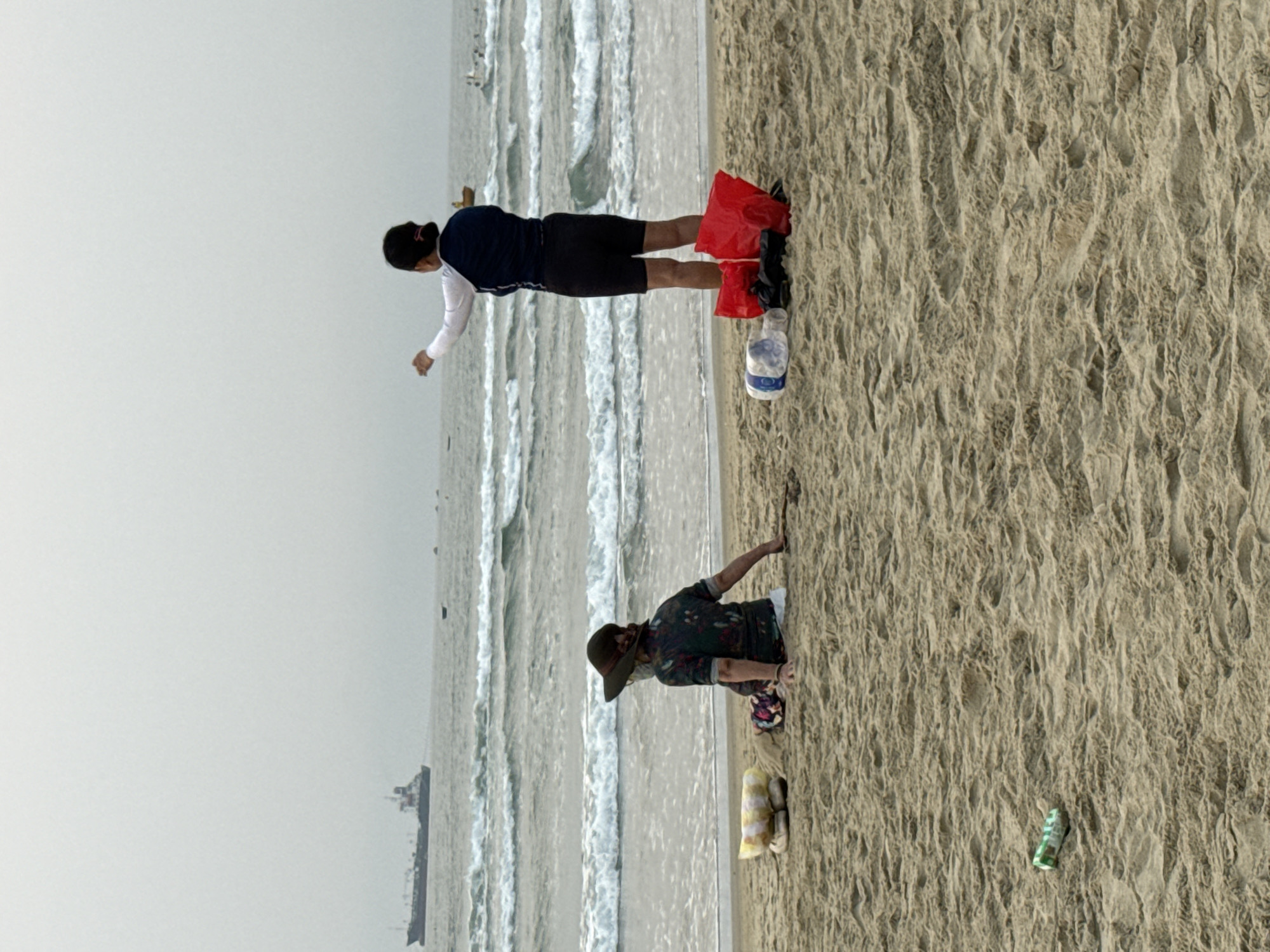 The image depicts two people on a sandy beach next to a body of water, likely the East Sea given Đà Nẵng's location. The sky is overcast.
Person 1, closer to the camera, is kneeling on the sand and reaching out toward the water's edge. They are wearing a dark-colored shirt with a possible floral or camouflage pattern, long, patterned pants, and a wide-brimmed hat.
Person 2, further away from the camera, is standing on the sand closer to the water. They are wearing a dark blue/black top with lighter-colored sleeves, dark shorts, and have red bags near their feet along with a plastic water bottle. They are reaching out toward the water's edge.
In the water, there are visible waves and a ship further out. The sand shows footprints and other disturbances. Trash, including a drink can and a plastic bag, is scattered on the sand.