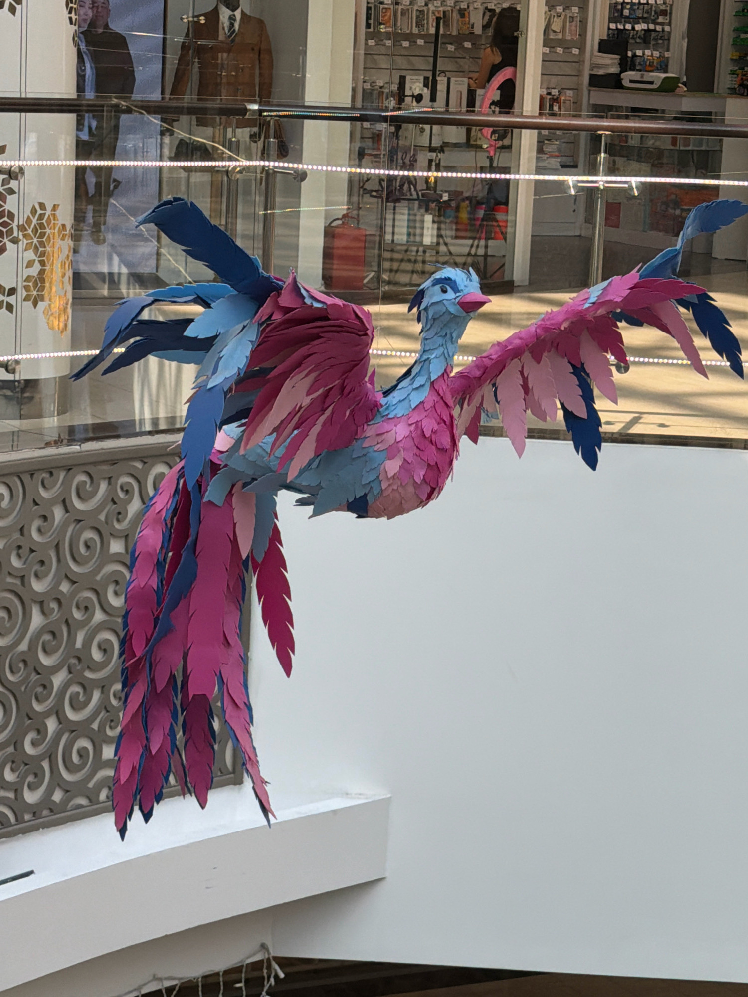 A colorful, artificial bird sculpture is suspended by wires in what appears to be a shopping mall. The sculpture is predominantly pink and blue, with numerous layered paper feathers forming its wings, tail, and body. The bird's head is facing left, with a light blue beak and eyes. In the background, through a glass partition, mannequins are displayed in store windows, one wearing a brown jacket and another a dark jacket. A woman with dark hair wearing a pink strap top is visible in the background, standing near a store counter with shelves of products. The environment suggests an indoor commercial space with lighting fixtures and architectural elements like railings and decorative wall patterns. The city of Pavlodar, Kazakhstan is not directly identifiable from the visual elements in the image.