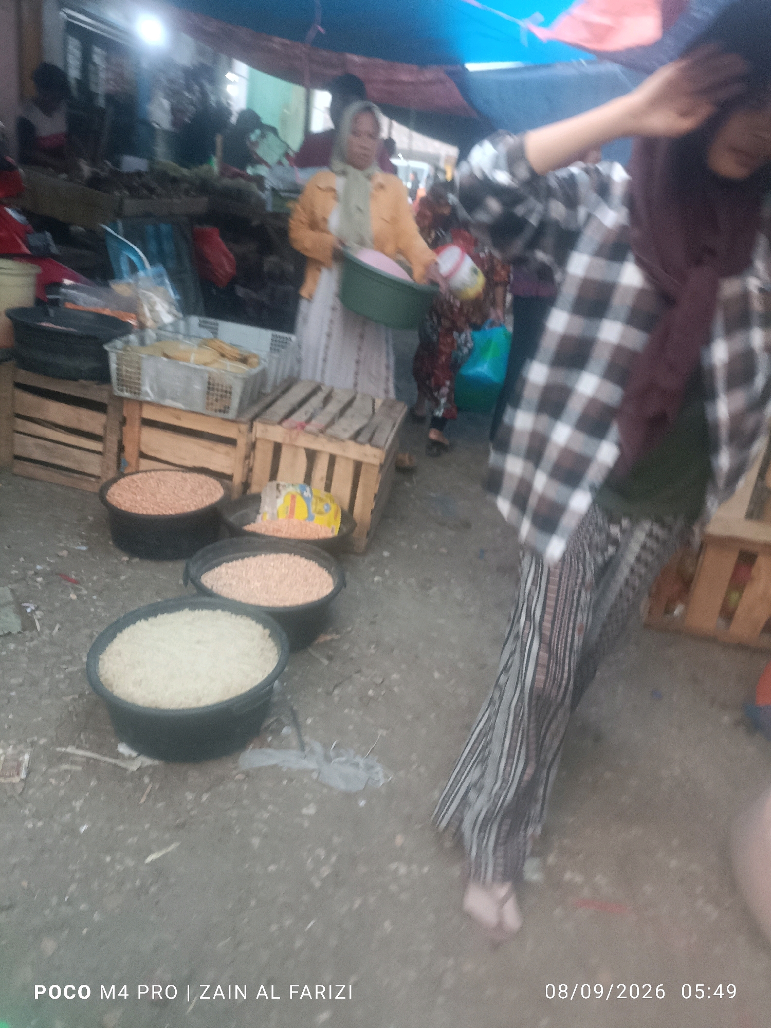 In Arosbaya, Indonesia, a street market scene unfolds. The foreground displays three black basins filled with granular food items: the closest one holds white rice, the middle one contains a lighter-colored grain, and the furthest basin is filled with a reddish-brown lentil-like food. A fourth basin behind these, partially obscured, also contains a similar reddish-brown grain, with a yellow and red bag visible inside it. Wooden crates are stacked and arranged around these basins, some empty and others supporting white plastic baskets containing what appear to be dried goods. A person in the middle ground, wearing a light-colored headscarf and an orange jacket, holds a green basin. Another individual in the right-middle ground is seen from the back, wearing a checkered shirt and patterned trousers. The background is blurred, showing more market stalls and people. The ground surface is a rough, dusty texture. The timestamp "08/09/2026 05:49" is overlaid on the bottom right of the image, along with "POCO M4 PRO | ZAIN AL FARIZI" on the bottom left.