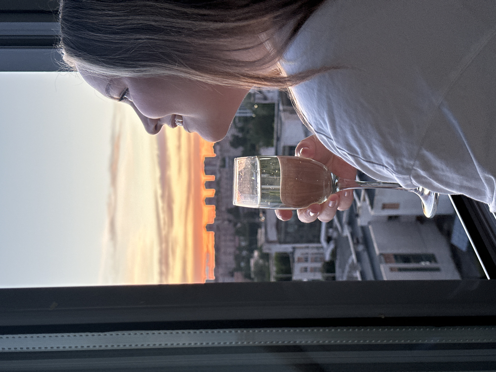 A side-view shot of a woman holding a wine glass in front of a window. The woman has light brown hair and a white shirt. A city and sunset are visible behind her through the window. The city in the background is possibly Izhevsk, Russia. The image appears to be taken indoors near a window. The woman is looking down towards the glass in her hand.