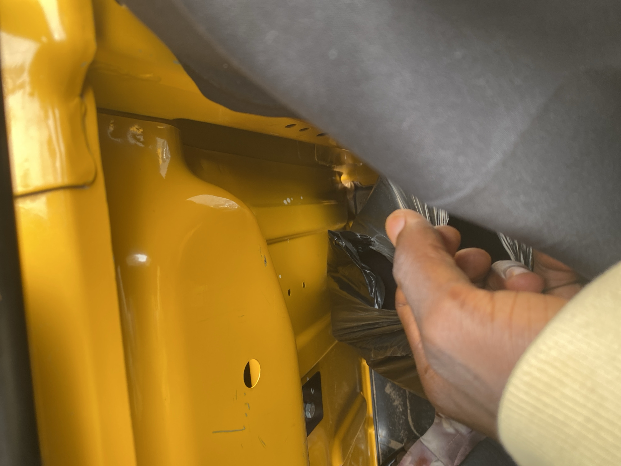 A close-up, overhead shot shows a person's hands reaching into a bright yellow interior, likely a vehicle. The yellow surface has a glossy finish and features several circular indentations and small holes. A dark grey material, possibly a fabric or plastic covering, is visible along the top edge of the frame, partially obscuring the yellow interior. The person's hands are manipulating a black, crinkled material, possibly plastic wrapping, which is tucked into the yellow space. The person is wearing a light yellow long-sleeved garment. The lighting suggests it is daytime, and the scene appears to be in a sheltered or enclosed area. No text is visible in the image. The context provided places this scene in Jalingo, Nigeria.