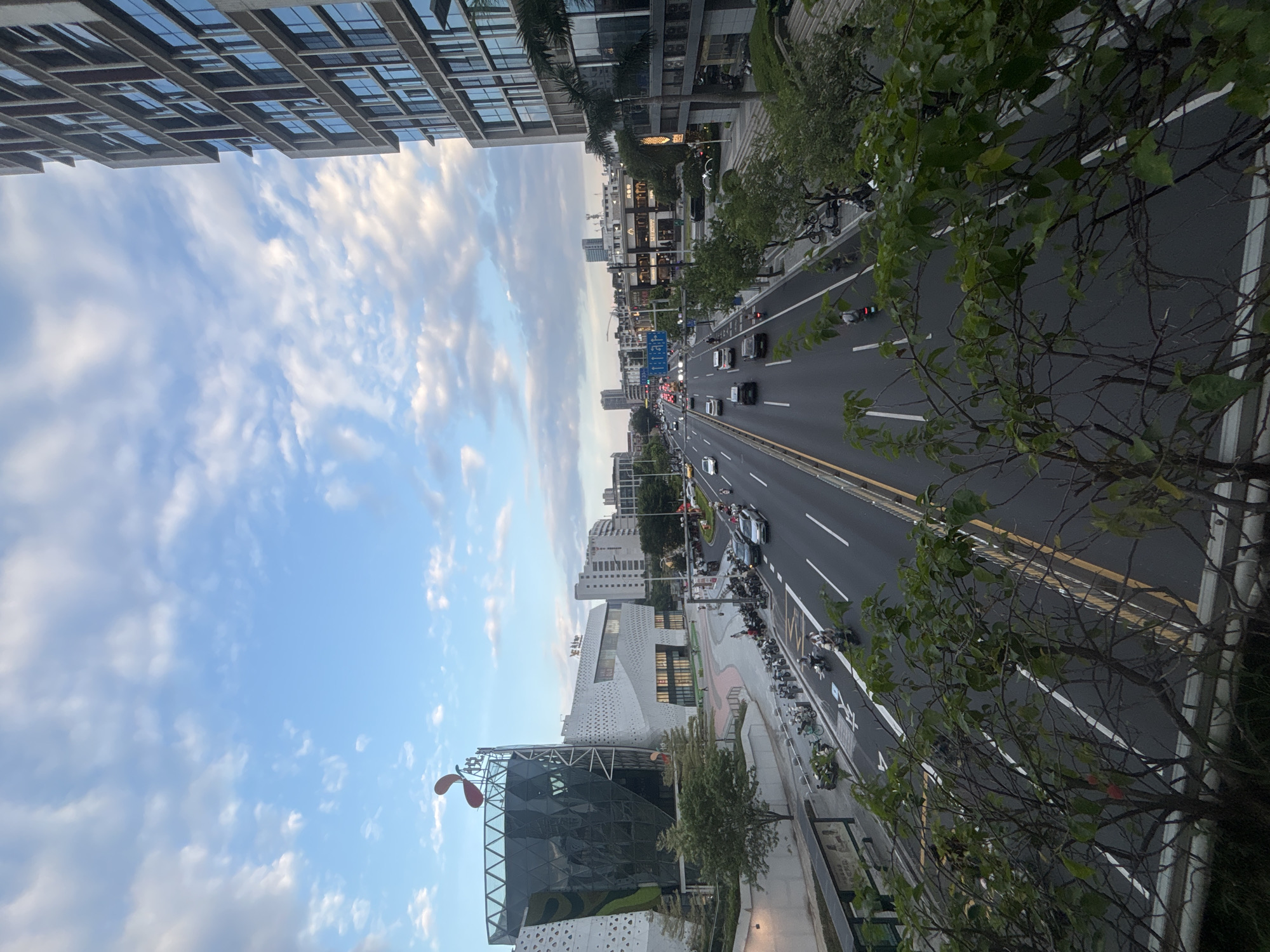The image captures an urban street scene with traffic, buildings, and vegetation. A multi-lane road with vehicles, including cars and motorcycles, runs through the center of the frame. On the left side of the road, a modern, angular building with a glass facade and white patterned walls stands prominently. A red, abstract sculpture is visible atop this building. Further down the street, other buildings of varying heights and architectural styles line the road, some appearing to be commercial establishments with signage. On the right side of the street, a tall, modern building with a grid-like facade of windows and concrete is depicted. Trees and foliage are present along the sidewalks and in planters, with some branches obscuring the foreground. The sky above is partly cloudy, with a mix of blue sky and white clouds. The overall impression is of a bustling city environment, likely during the daytime or early evening. The location is identified as Liwan District, China.
