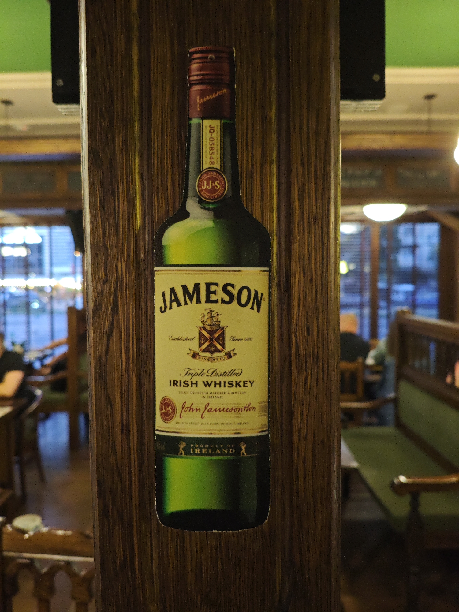 A Jameson Irish Whiskey bottle label is attached to a wooden beam in what appears to be a dimly lit establishment. The label is green and yellow, with prominent text reading "JAMESON IRISH WHISKEY" and "John Jameson". The background shows a blurred interior with tables, chairs, and patrons, suggesting a bar or restaurant setting. There is no visual information in the image to indicate its location in Kyiv, Ukraine.