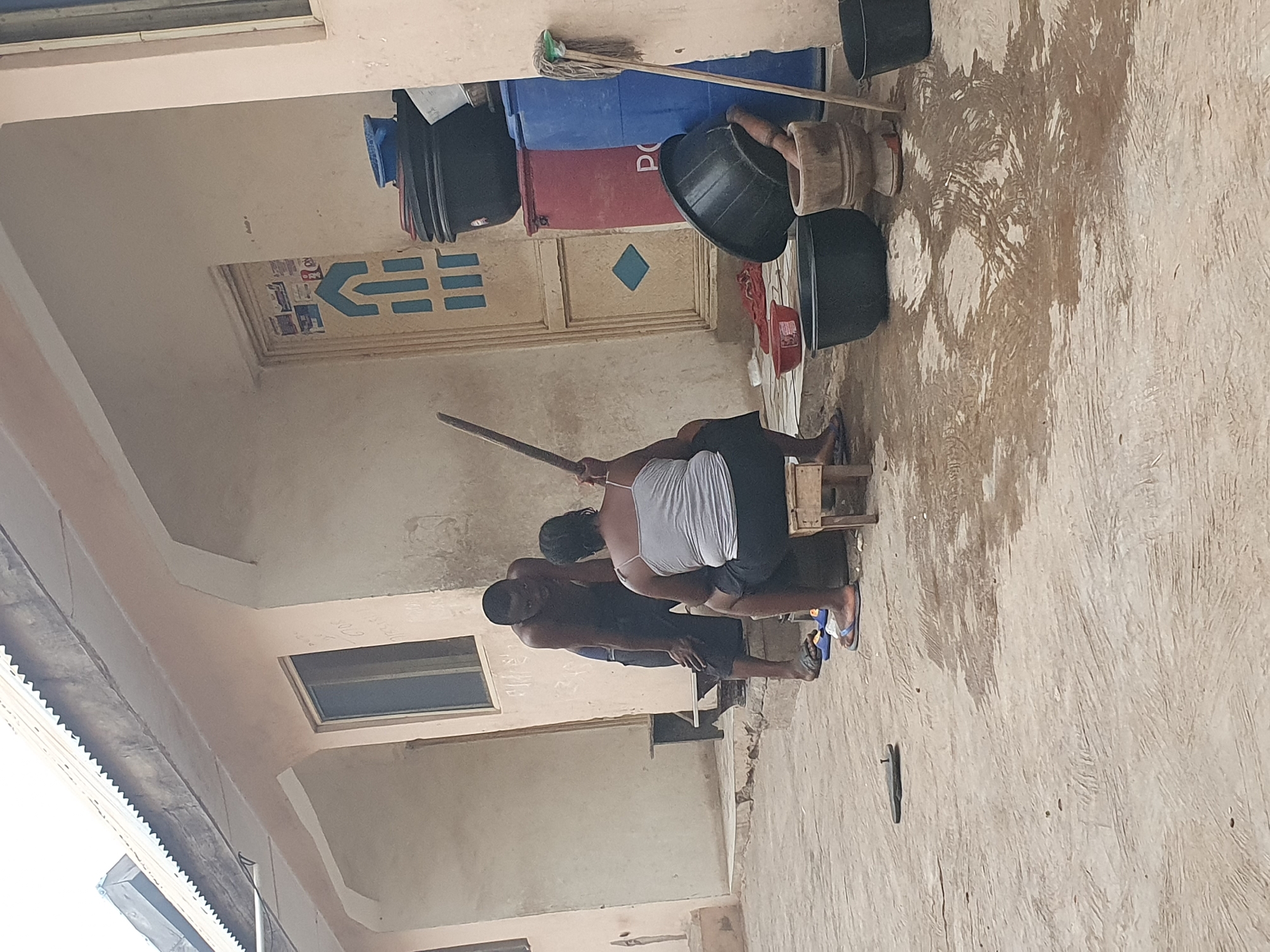 Two people sit on a wooden stool outside a building in Lokoja, Nigeria. One person is holding a long wooden stick. The building is painted beige and has a door with blue decorations. Several black buckets, a blue barrel, and a red barrel are stacked against the wall. A mop leans against the barrels. The ground is light-colored and textured. The building's roofline is visible on the left. A window is present in the building wall.