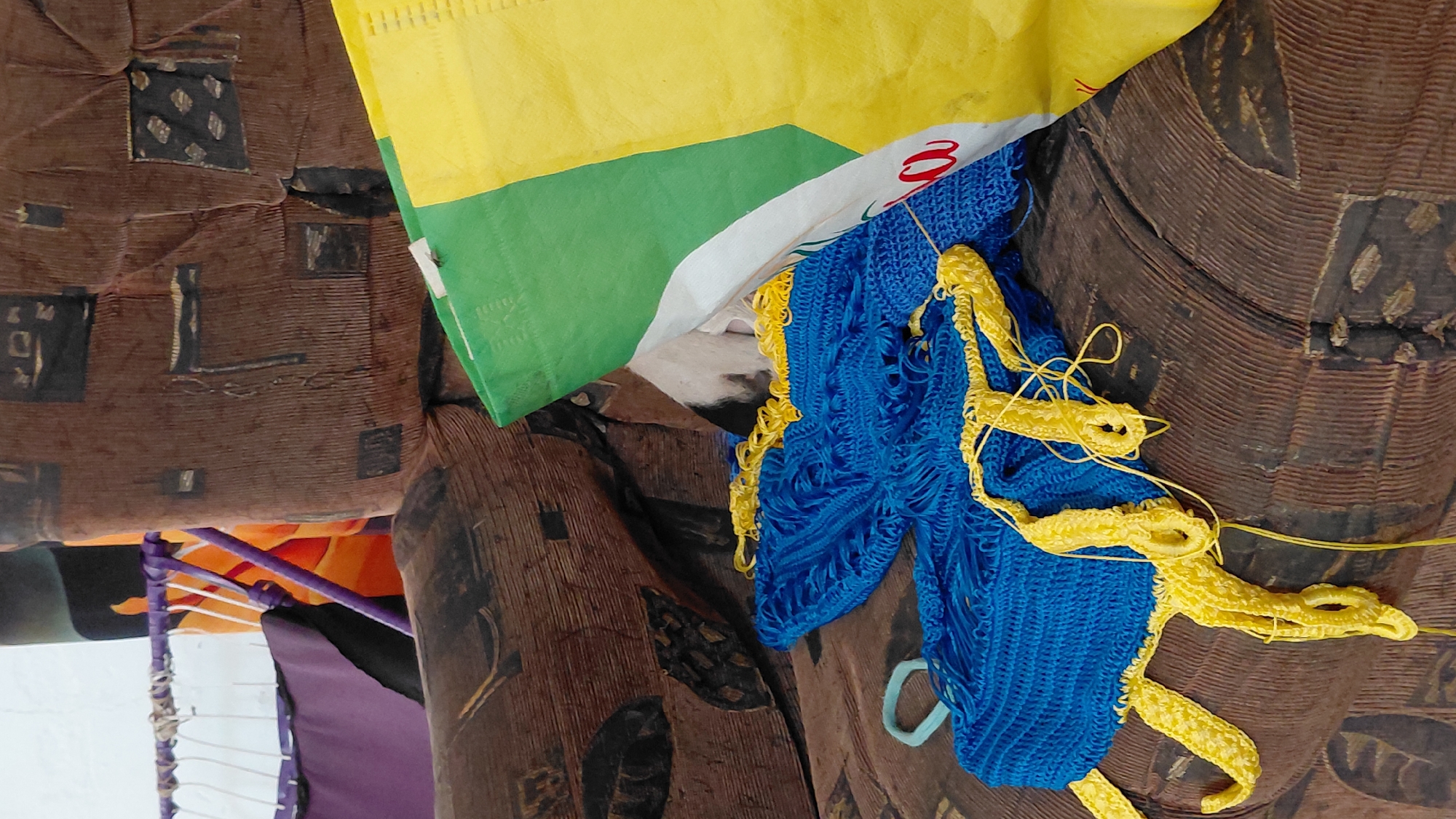 A close-up, eye-level shot shows a portion of a brown corduroy sofa. On the sofa, a blue crocheted garment with yellow trim is laid out, its fringe and unfinished edges visible. To the right of the garment, a large green and yellow woven bag is partially covering a small white and black dog, which is peeking out from under the bag. The background shows parts of a purple and orange draped fabric and a white wall. The lighting is natural and diffused.