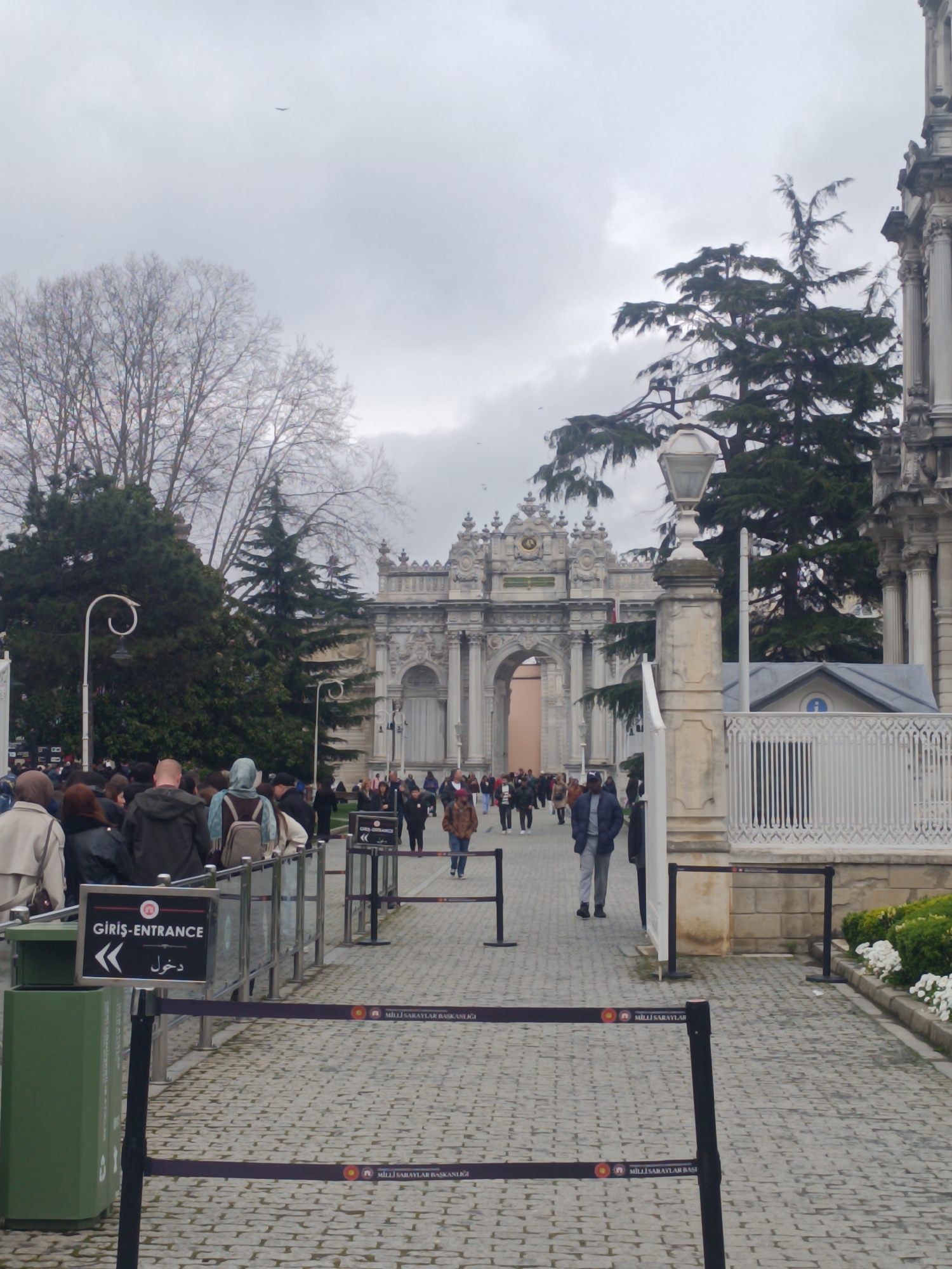 This eye-level, full outdoor shot depicts a paved area in the Beşiktaş district of Istanbul, Turkey, leading toward a grand archway building in the distance. The sky is overcast.
In the foreground, crowd control barriers form queues, with people waiting in line. A green trash receptacle sits on the left. The sign "Giriş-Entrance" is visible.
Beyond the queuing area, people are walking in both directions along the paved path. A large, ornate archway structure stands in the distance. Bare trees and evergreen trees are present. To the right, part of an architectural structure with columns and a small building with a white fence are visible. A lamp post stands to the right of the path.