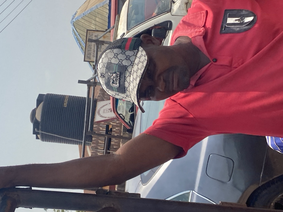 An adult male is partially visible in the foreground, wearing a red polo shirt with a dark emblem on the left chest and a white and grey patterned trucker cap featuring a "GUCCI" logo and green/red stripe detail. His right arm is raised, grasping a vertical metallic pole or frame to his left. He has a short beard or stubble and a neutral expression, looking slightly downwards.
In the mid-ground, a dark grey sedan is parked to the left, partially obscured by the man and the pole. To the right of the man, a white vehicle (possibly a van) is parked. Further back, between these vehicles, a red vehicle is visible.
The background features two large, black, ribbed plastic water tanks, one bearing the text "AMURUMU" or similar. Behind the tanks and vehicles, a wooden sign clearly reads "AMURO MGBOM COMMUNITY PRIMARY SCHOOL," establishing the location in Afikpo, Nigeria, near this institution. A light-colored, possibly corrugated metal, pyramidal roof is also visible behind the sign. The sky is clear, indicating daytime.