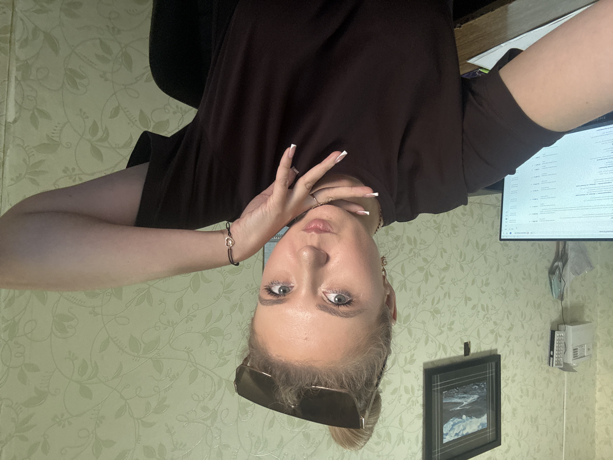 The image shows a young woman with blonde hair, wearing sunglasses on her head and a dark brown t-shirt. She is posing for a selfie, with her hand near her face. Her nails are painted with a French manicure. She has light blue eyes and is wearing a bracelet with a heart-shaped charm. The background appears to be a room with light green wallpaper featuring a floral pattern. To the right of the woman, a computer monitor is visible, displaying text that is too small to read. There is also a framed picture on the wall and some electronic devices. The image is rotated 90 degrees clockwise, so the woman's head is at the bottom. The lighting suggests it could be daytime, but the exact time is not discernible. The overall impression is of a casual, indoor setting.