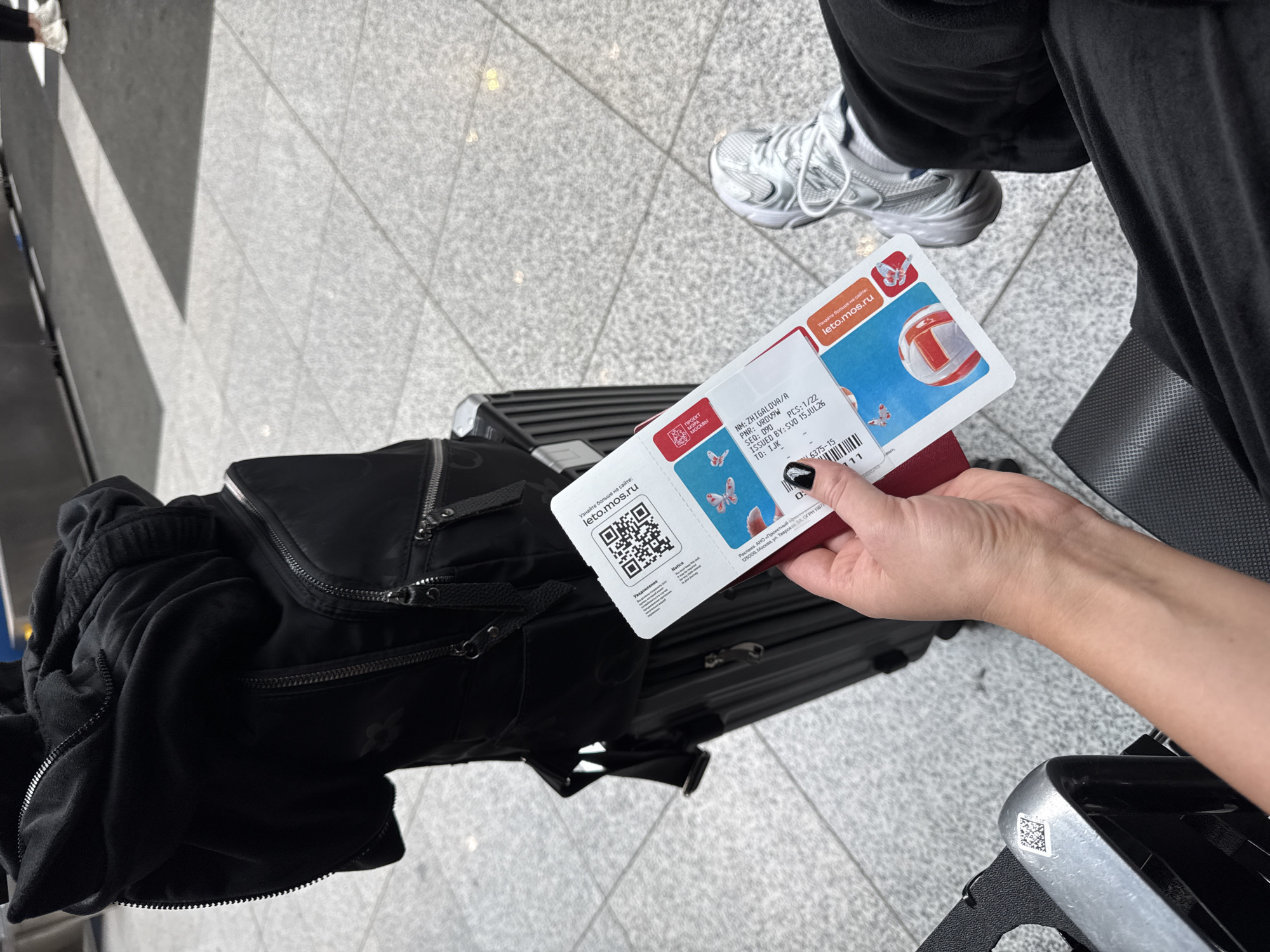An overhead shot shows a person's hand holding a boarding pass and a passport in an airport terminal. The boarding pass is white with colorful graphics of butterflies and a volleyball, and the text "leto.mos.ru" is visible. The passport is red. A black backpack and a black suitcase are visible behind the hand. The person is wearing a black hooded sweatshirt and black pants, and white sneakers. The floor is tiled with a gray and white pattern, and some white lines indicate seating areas. The background is blurred, but appears to be the interior of an airport. The scene is brightly lit by overhead lights.