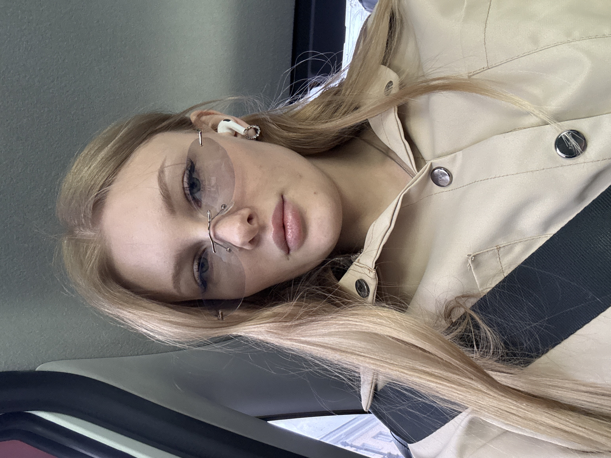 A young Caucasian woman with long, blonde hair is the primary subject of the image. She is wearing frameless glasses and what appears to be a light beige jacket or coat. She also has an earpiece in her left ear. A black seatbelt is visible across her body.
The interior of a vehicle, likely a car, is discernible in the background, along with the car door and the outside seen through the window.
The location is unspecified, but based on the prompt's information, it is presumed to be Perm, Russia.