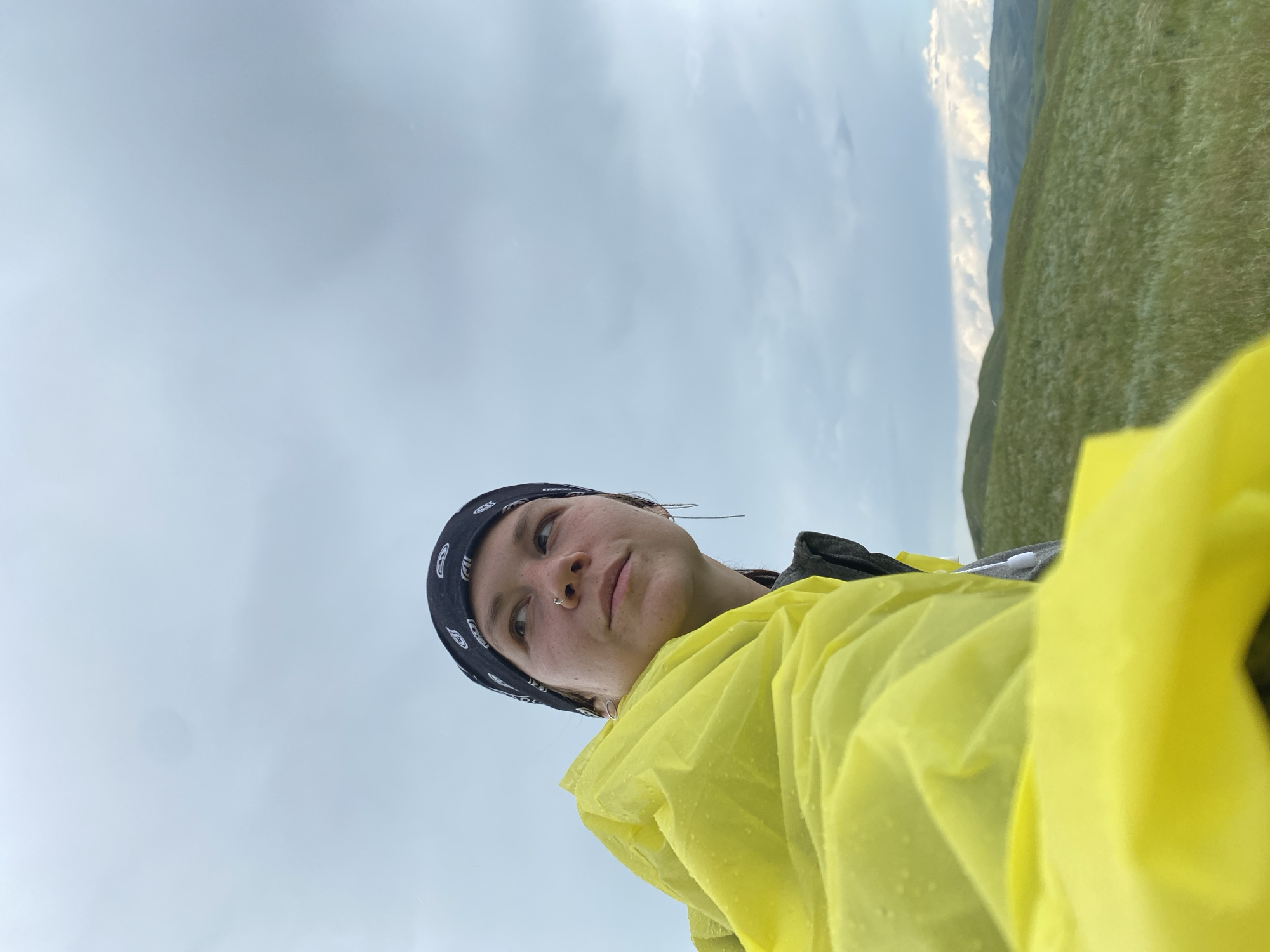A young person with a nose ring and a black bandana is looking up and to the right, partially covered by a bright yellow raincoat with water droplets on it. They are outdoors, in a grassy, hilly landscape under a cloudy sky. The image appears to be a selfie taken in a natural environment, possibly on a hike or outdoor excursion. The background consists of rolling green hills and a vast expanse of sky filled with grey and white clouds, suggesting the possibility of inclement weather. The location is identified as the city of Rykulovo, Russia.