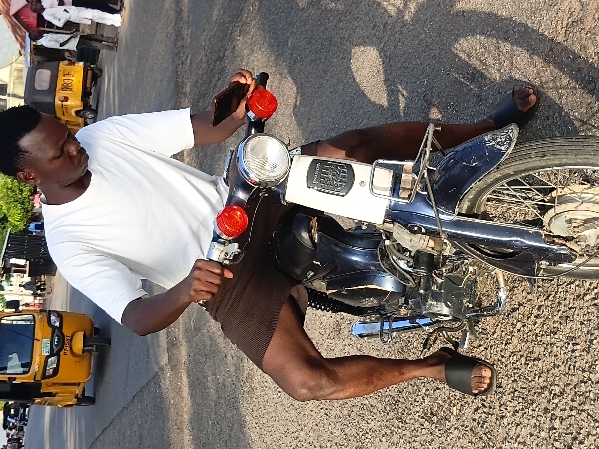 The image depicts a young man sitting on a dark blue and white motorcycle on a paved road in Jalingo, Nigeria. He is wearing a white t-shirt and brown shorts. The man is holding a dark smartphone in his right hand. The motorcycle has a white front panel with a "HONDA" logo and a chrome headlight. Red turn signals are visible on the handlebars. In the background, a yellow auto-rickshaw is parked on the side of the road, along with several people. The ground is covered in asphalt with scattered gravel. The overall lighting suggests it is daytime, with shadows indicating the sun is relatively low in the sky.