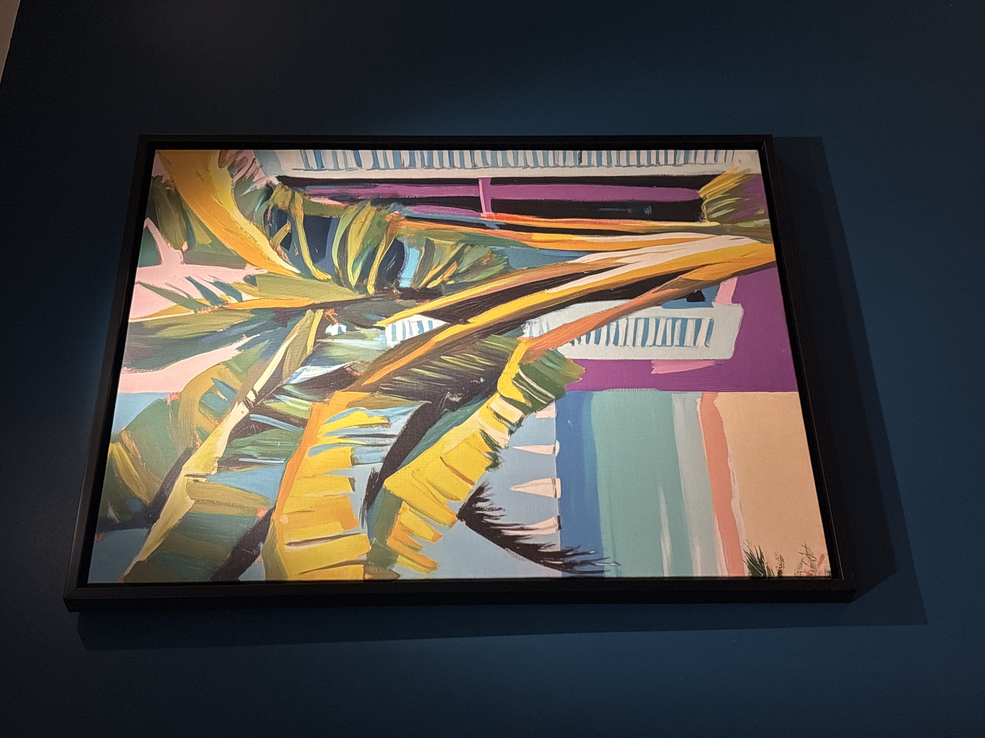 The image shows a framed painting with a black frame, hanging on a dark blue wall. The painting is an impressionistic depiction of a tropical scene. A large banana leaf plant dominates the foreground, with bright yellow and green leaves. To the right, a purple building with white shutters is visible. In the background, a beach scene with pale orange sand and turquoise water is depicted, with three white sailboats on the horizon under a light blue sky. The painting is positioned at an angle, with a shadow cast on the wall below and to the left. The location of the artwork is not specified in the image, and therefore cannot be described as being in Amman, Jordan.