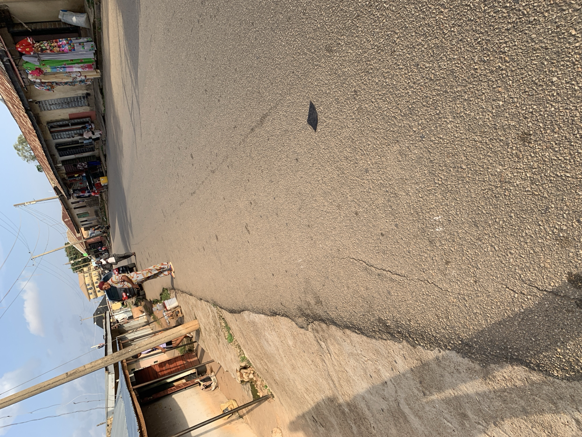 This media file depicts a street scene in Jos, Nigeria. The image is taken from a slightly elevated perspective, looking down at an asphalt road. On the left side of the road, there are buildings with shops and market stalls. These stalls display various goods, including colorful fabrics and other items. Some people are visible in front of the shops, and one person appears to be a vendor.
Further down the street, a dark-colored car is parked, and a few individuals are standing near it. The architecture of the buildings suggests a tropical or sub-tropical climate. There are utility poles with electrical wires running along the street, indicating a developed infrastructure.
The road itself shows signs of wear, with some cracks and rough patches. A small, dark object is present on the road surface in the middle-right of the frame. The lighting suggests it is daytime, with the sun casting shadows. The sky is visible in the upper left corner of the image and appears clear, indicating fair weather. The overall impression is of a bustling market or commercial area on a typical day. No visible text is discernible in the image.