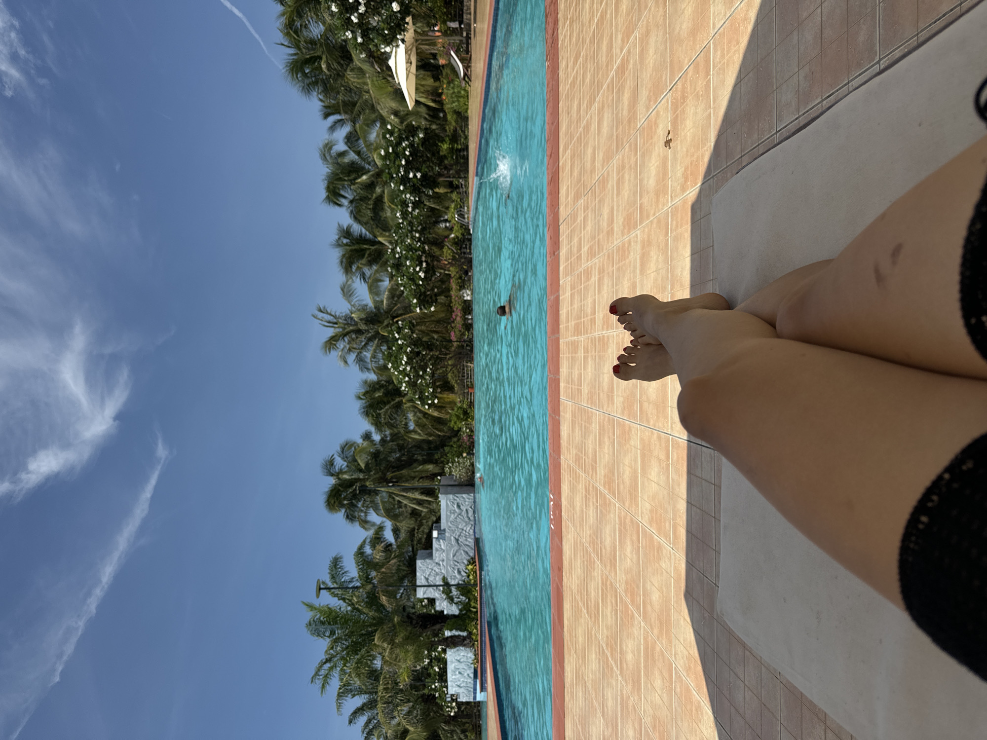 The image depicts a person lying on a lounge chair by a swimming pool. The person's legs and feet are in the foreground, with painted red toenails visible. The pool is filled with blue water and appears to be outdoors. Several people are swimming or wading in the pool. Palm trees and flowering bushes line the perimeter of the pool, creating a tropical atmosphere. The ground around the pool is tiled. In the background, there is a clear blue sky with some wispy clouds. The overall scene suggests a relaxing day at a resort or hotel in Phu Thuy Ward, Vietnam.