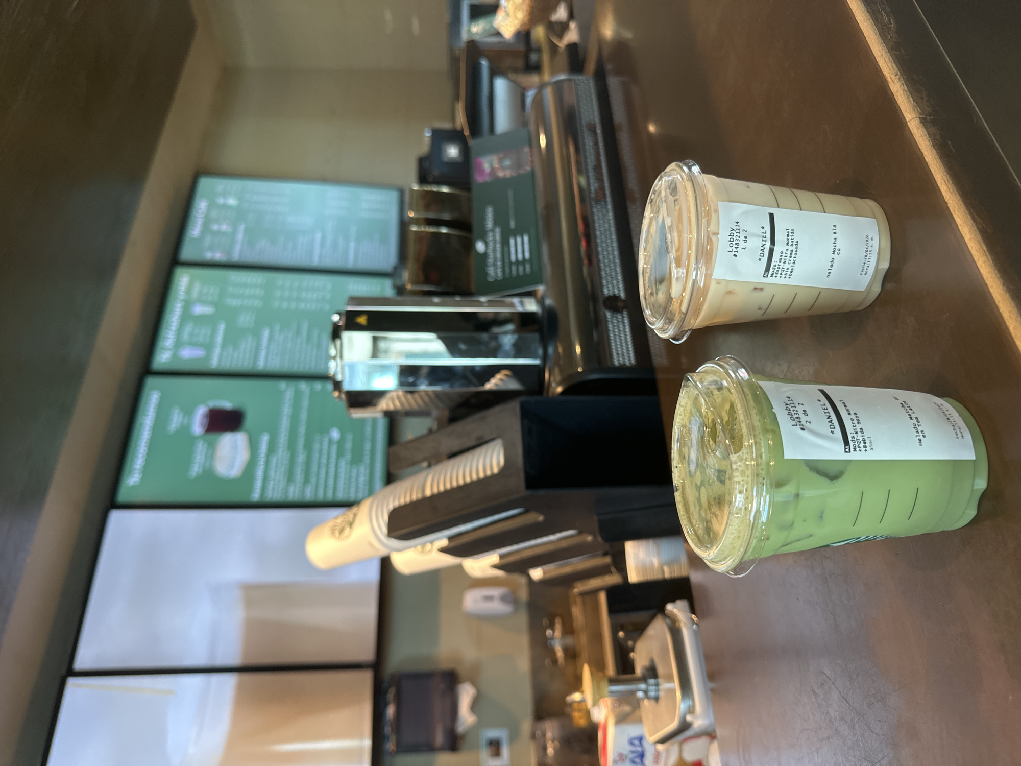 This image displays an indoor scene at a coffee shop, likely Starbucks, featuring two iced beverages on a dark brown counter. Both drinks are in clear plastic cups with domed lids and custom white labels.
The drink on the right, labeled "1 de 2," is light brown/beige. Its label indicates it is for "*DANIEL*", order #14832114. Modifications listed are "Espresso," "+POT-Nitro Normal," "+Sin crema batida" (no whipped cream), and "+Deslactosada" (lactose-free). The beverage is identified as "Helado Mocha Blaco" (White Mocha Ice Cream) and dated "28/04/20" at "11:15 am."
The second drink, labeled "2 de 2," is a light green color, indicative of a matcha beverage. This drink is also for "*DANIEL*" from order #14832114. Its modifications include "AL," "+POT-Nitro Normal," "+Bebida Soya" (soy milk), and "Sincl." It is specified as "Helado Matcha Green Tea" (Matcha Green Tea Ice Cream) with a date of "28/04/2026."
In the background, out-of-focus digital menu boards with green screens and white text are visible. Various pieces of coffee preparation equipment and a cup dispenser with white paper cups are partially seen behind the drinks. The scene is brightly lit with indoor artificial light, indicating daytime. The labels are in Spanish, aligning with the San Andrés Cholula, Mexico location context. No individuals are present in the frame, but the prepared drinks suggest an ongoing customer transaction.